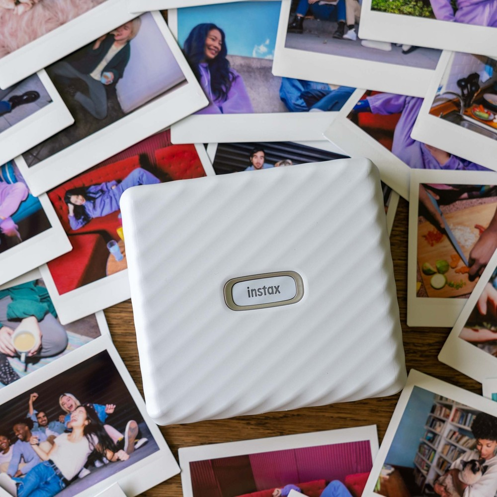 Ahşap üzerinde beyaz Instax yazıcı, insanlardan oluşan anlık fotoğraflarla çevrili.