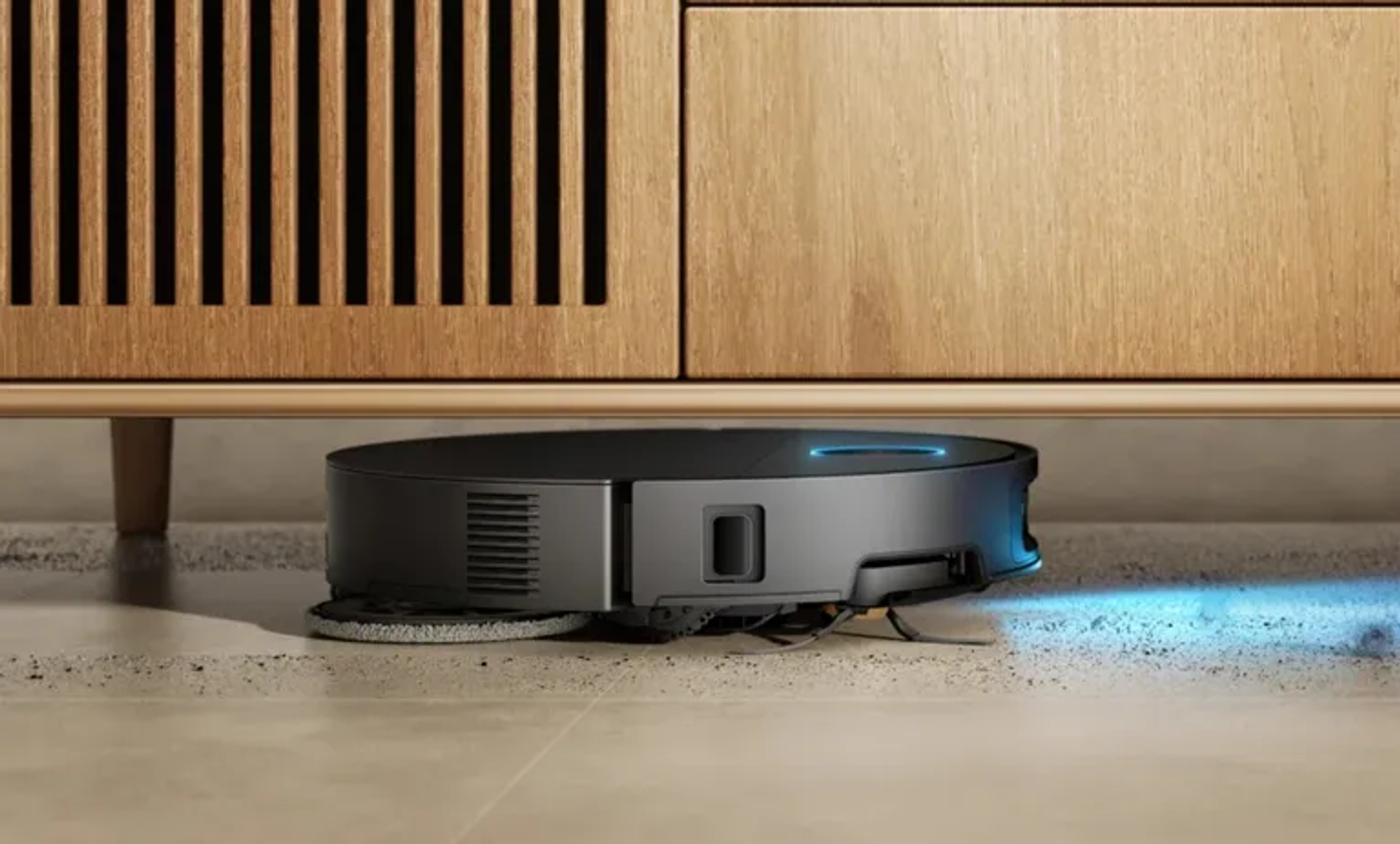 Un robot aspirador redondo y negro limpia una alfombra debajo de un mueble de madera.