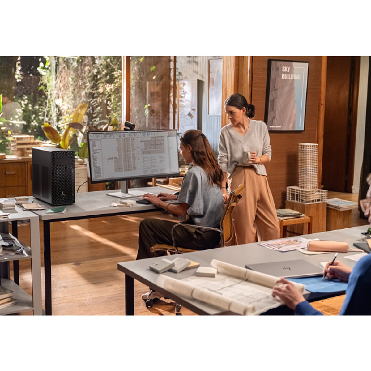 Dos mujeres trabajan en escritorios con computadoras y planos en una oficina luminosa.