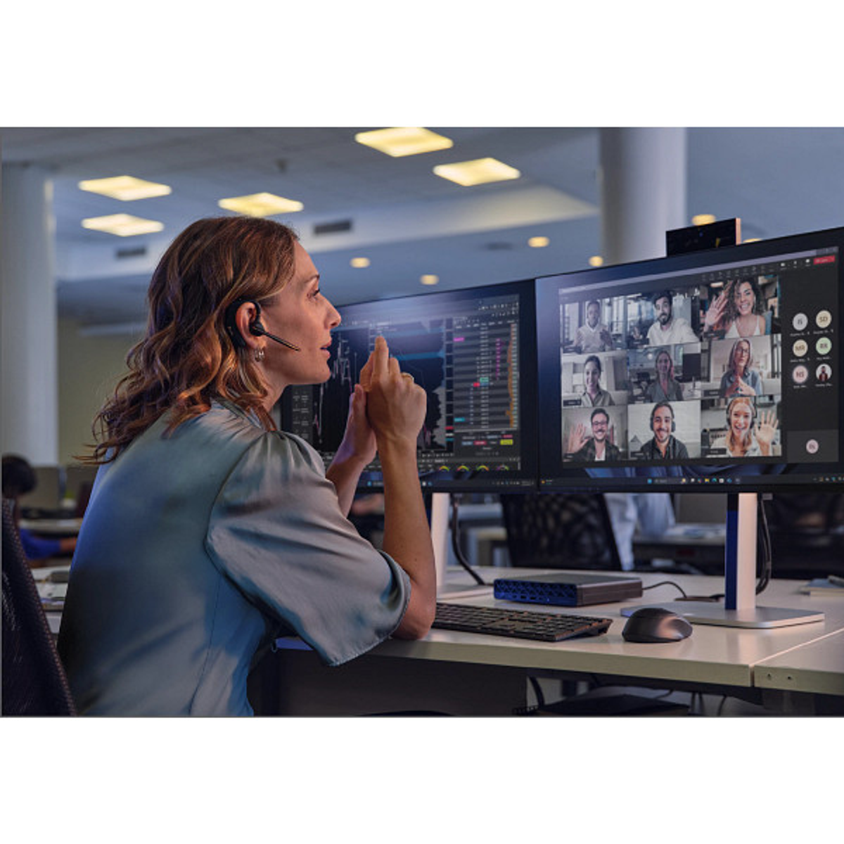 Donna con auricolare in videoconferenza con più partecipanti e dati di borsa sullo schermo.