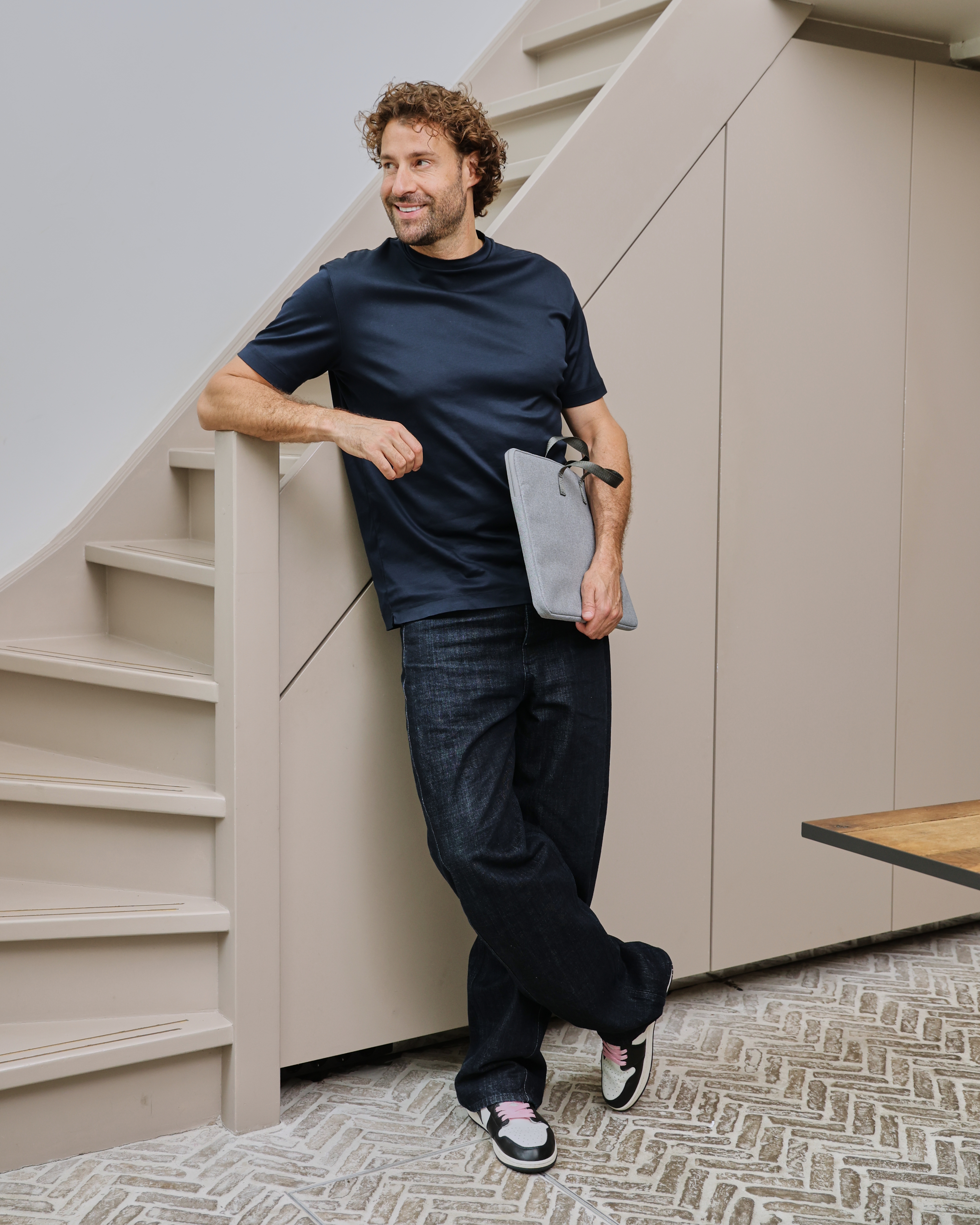 Een man met krullend haar leunt op een trap en houdt een laptoptas vast.