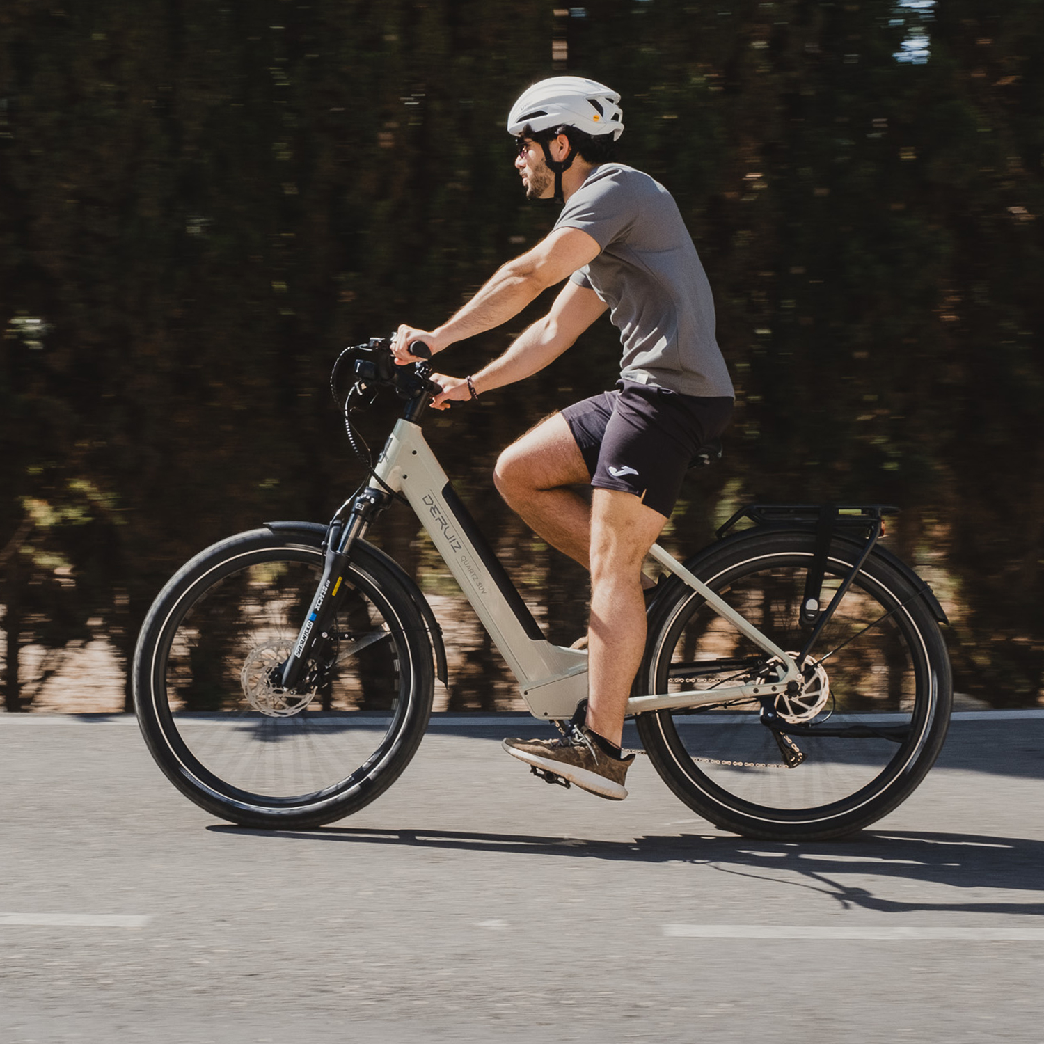 Mann fährt ein hellbeiges E-Bike auf einer Straße, Bäume im Hintergrund.