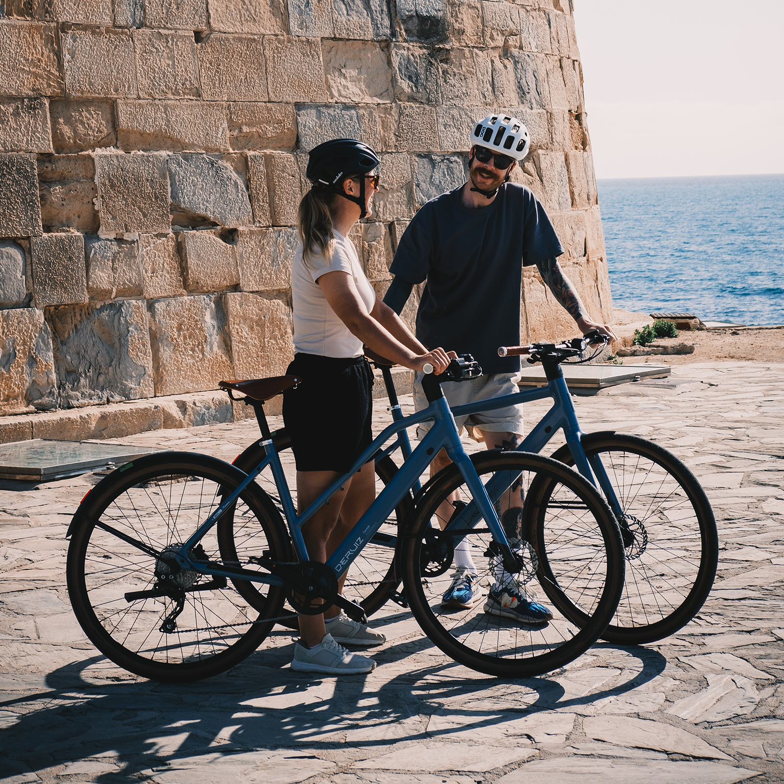 Ein Mann und eine Frau mit Fahrrädern stehen vor einer Steinmauer am Meer.