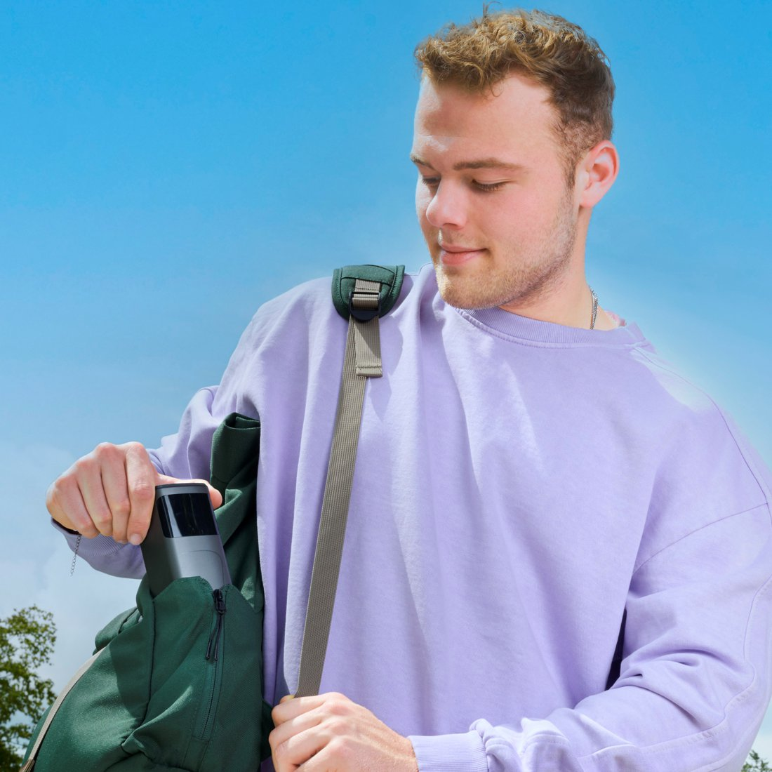 Ein Mann mit lila Pullover verstaut ein schwarzes Gerät in einem grünen Rucksack.