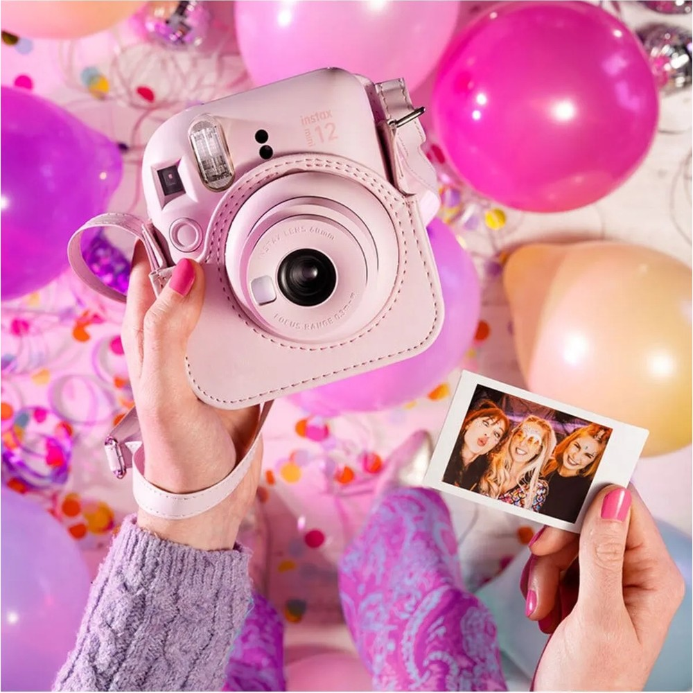 Pembe Instax kamera ellerle tutuluyor, renkli balonlar ve konfetilerle çevrili.