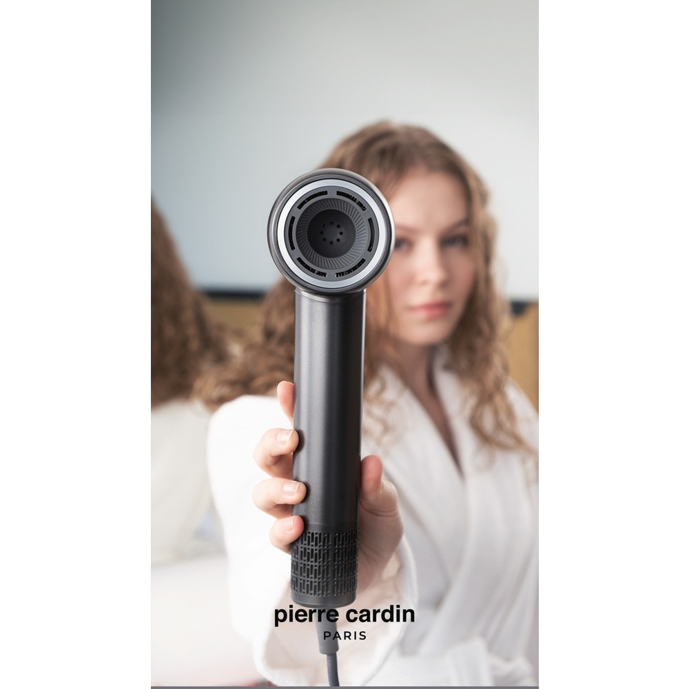 Beyaz bornozlu bir kişi kameraya siyah bir saç kurutma makinesi tutuyor. Pierre Cardin Paris logosu.
