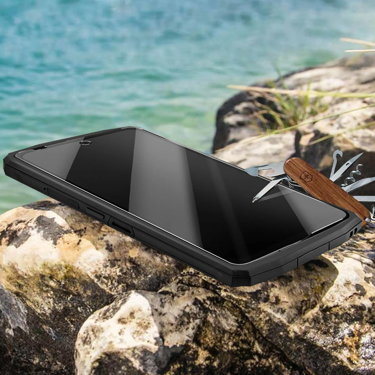 Uno smartphone poggia su rocce vicino al mare, con un coltellino svizzero accanto.