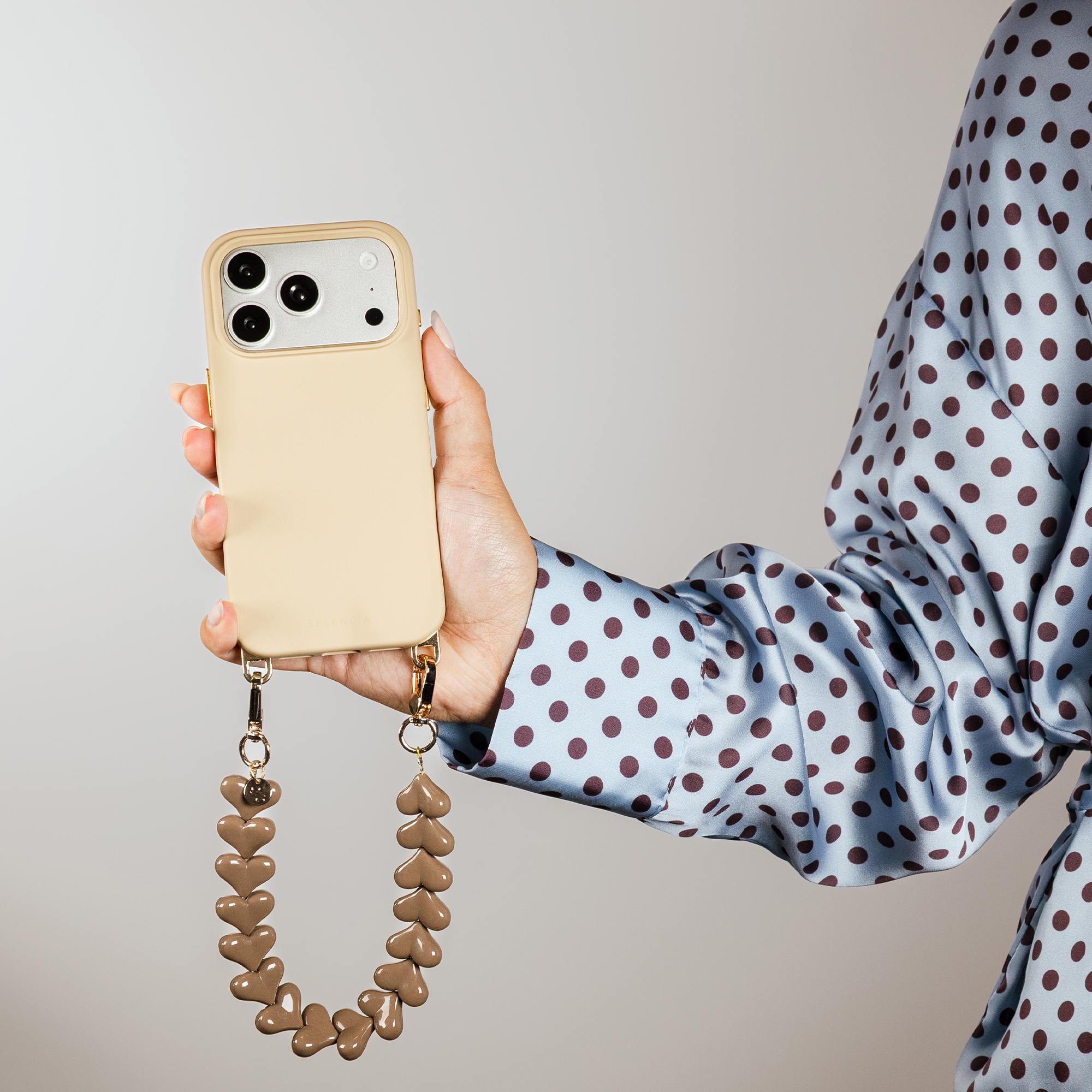 A hand holds a beige iPhone with a heart charm chain. Blue polka dot clothing.