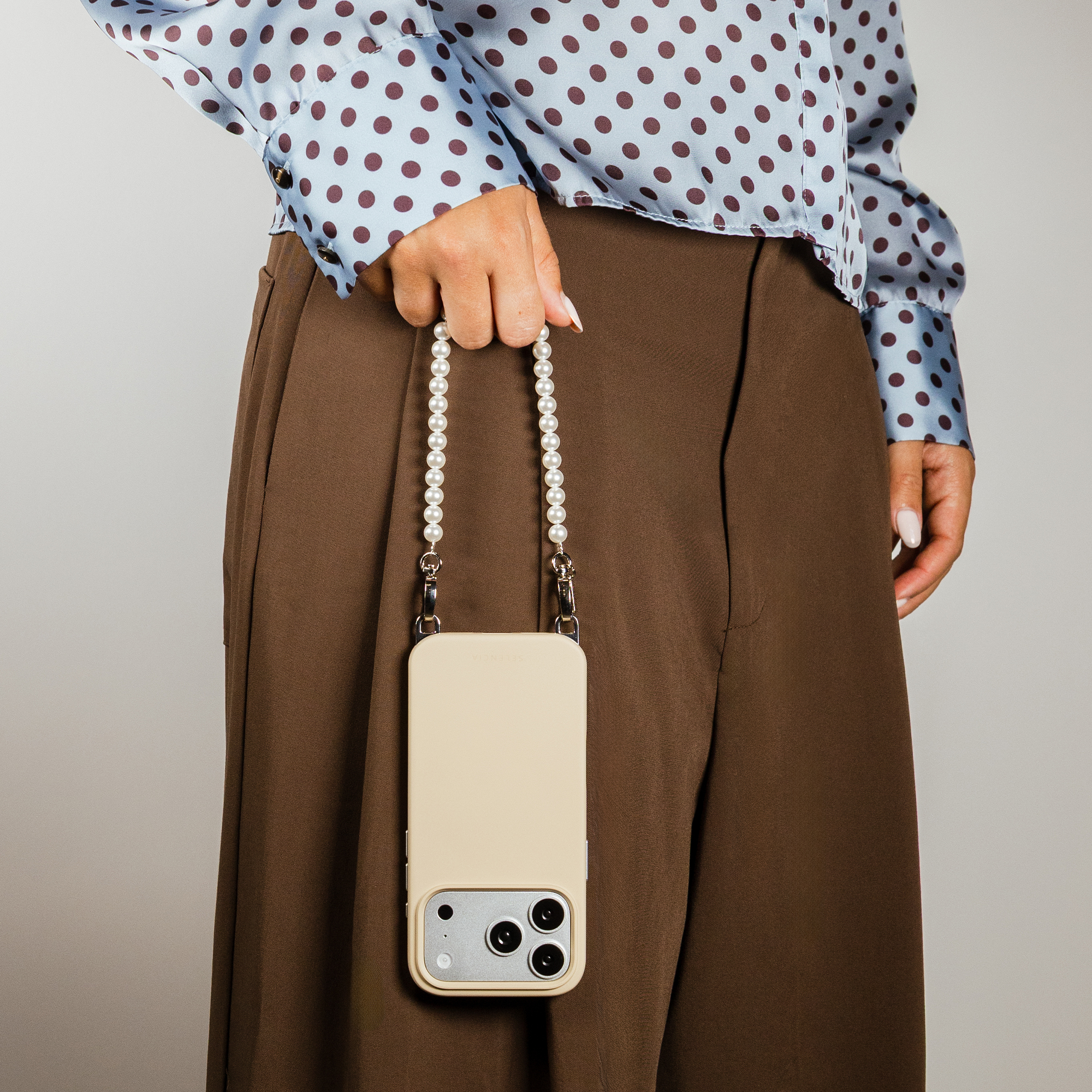 A person holds a beige phone case with a pearl strap. They wear a blue polka dot shirt and brown pants.