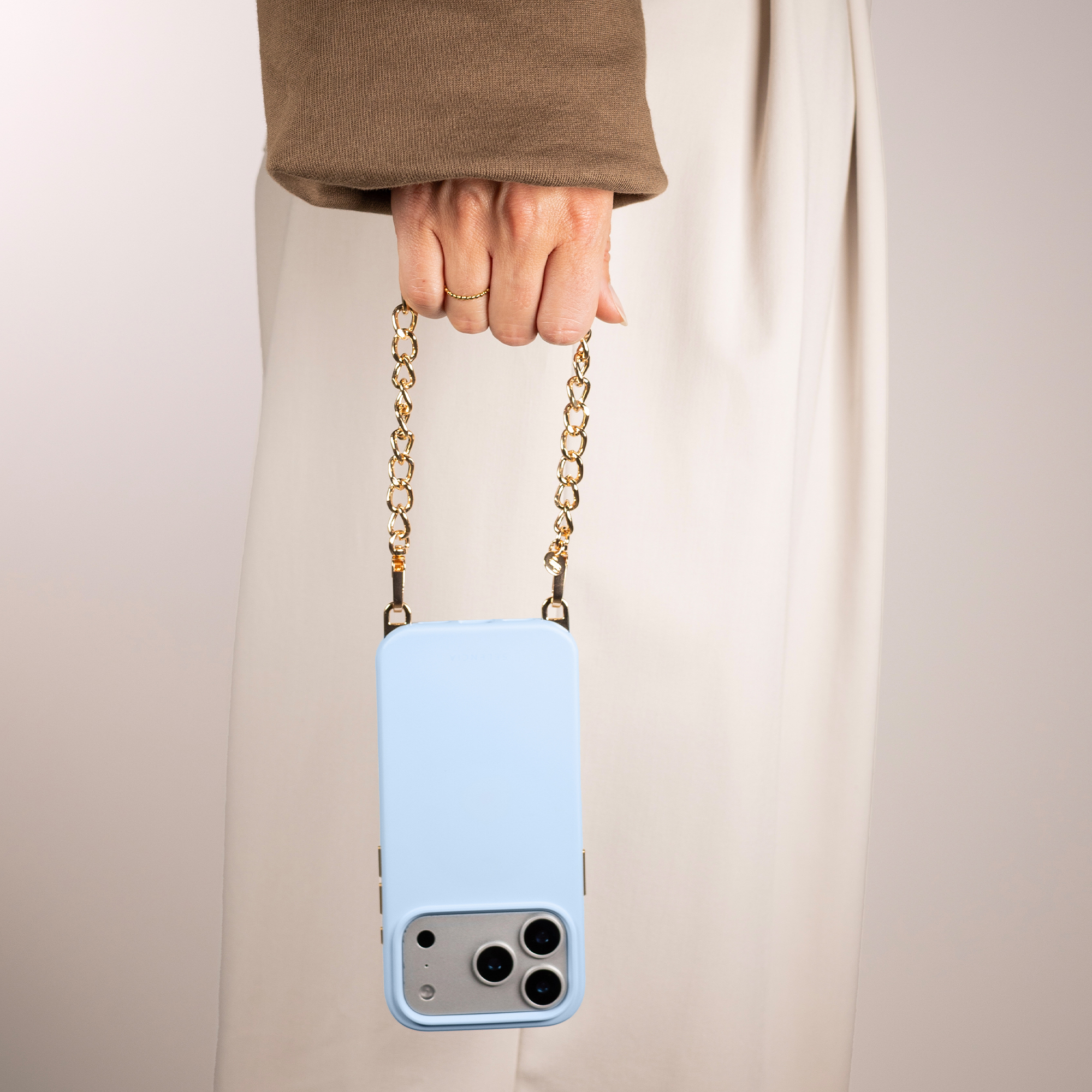 A hand holds a light blue phone with a gold chain.