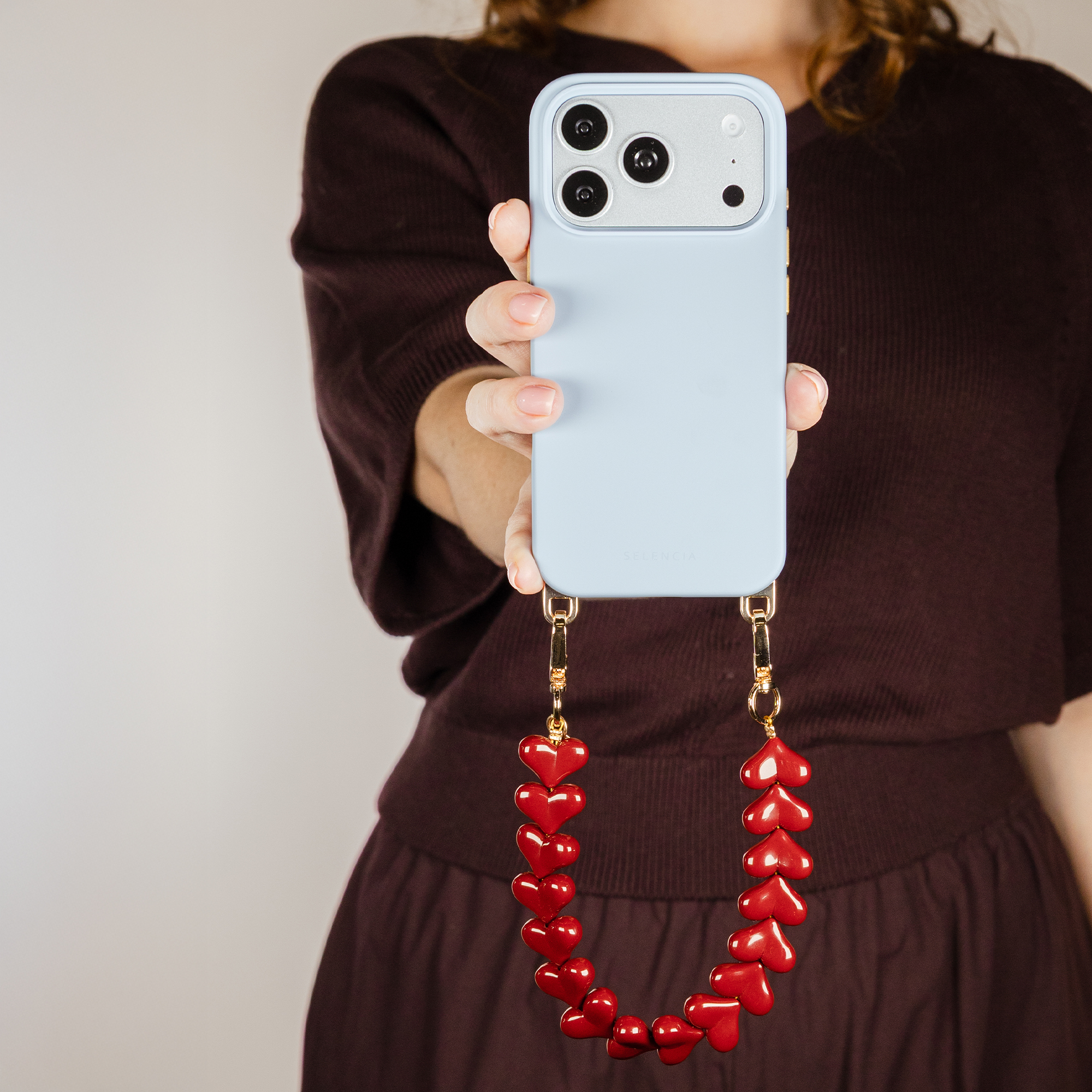 A hand holds a light blue phone with a red heart charm chain.