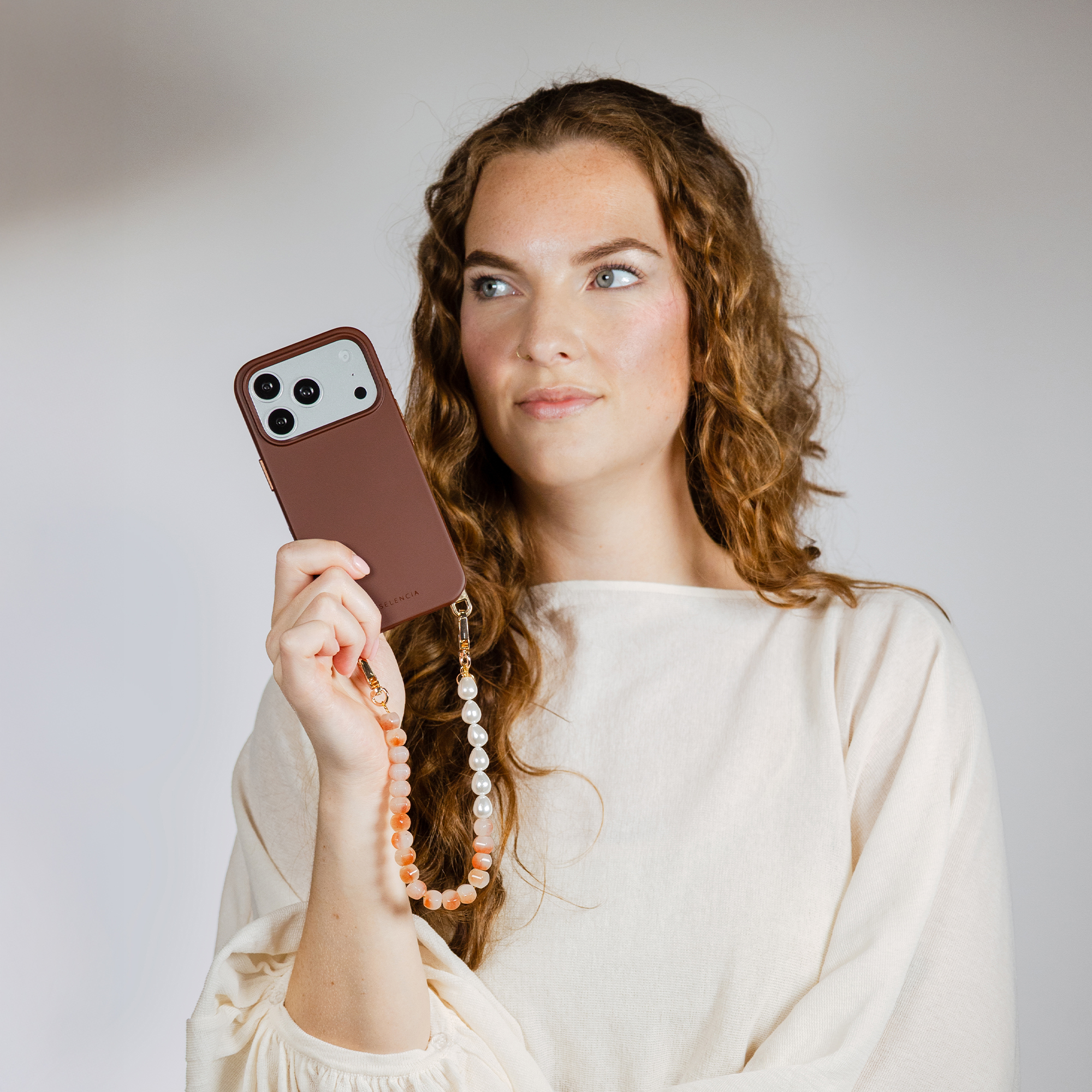 A woman with curly hair holds a brown phone with a pearl and stone charm.