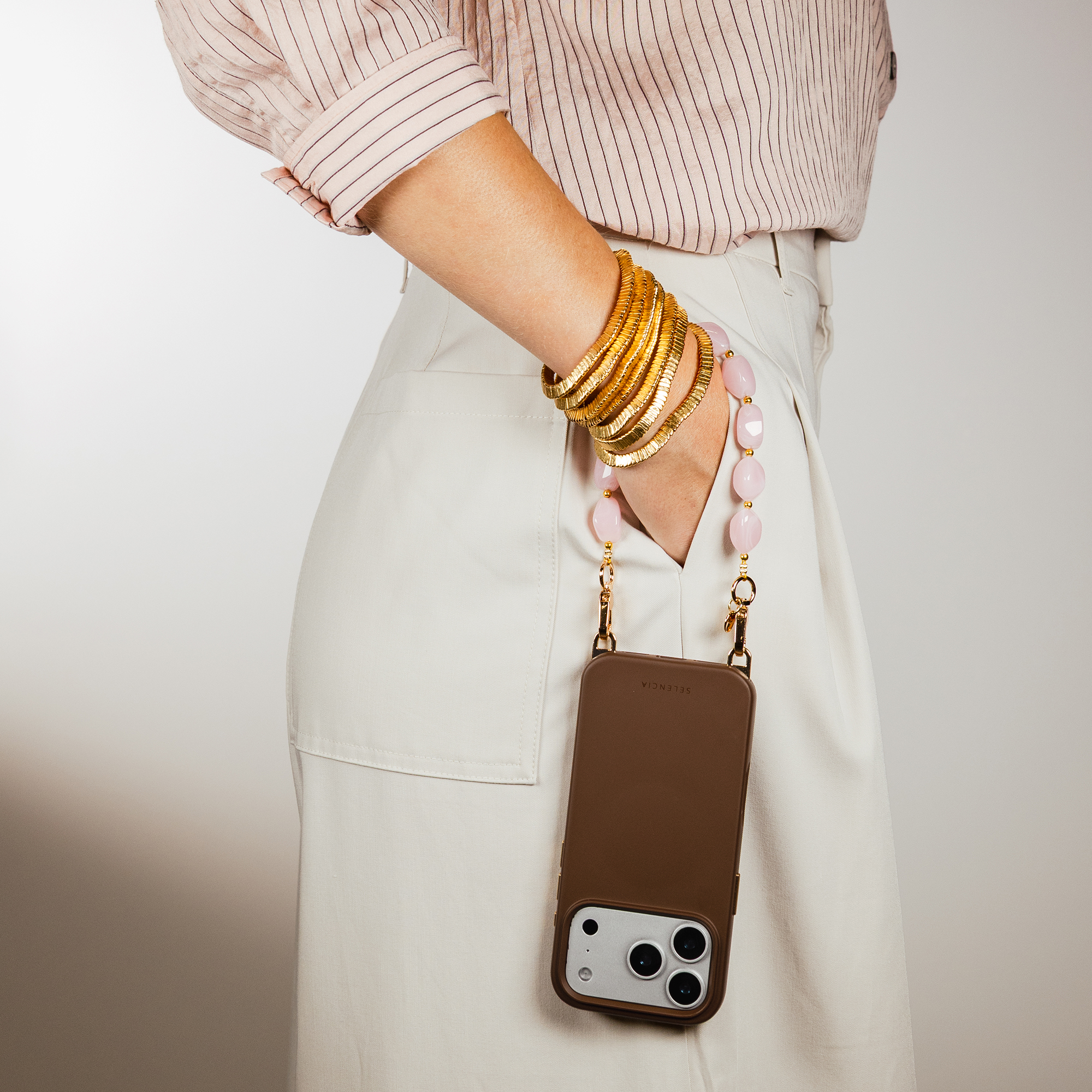 A woman wears a light brown top, beige skirt, gold bangles, and a brown phone case with pearls.