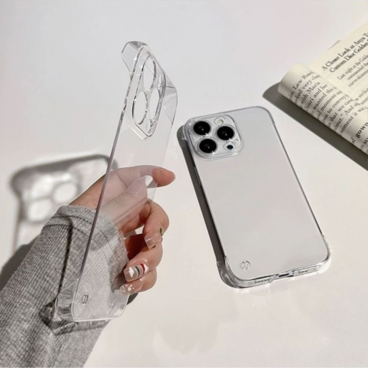 Eine Hand hält eine durchsichtige Handyhülle neben einem weißen Smartphone und einer gerollten Zeitung.