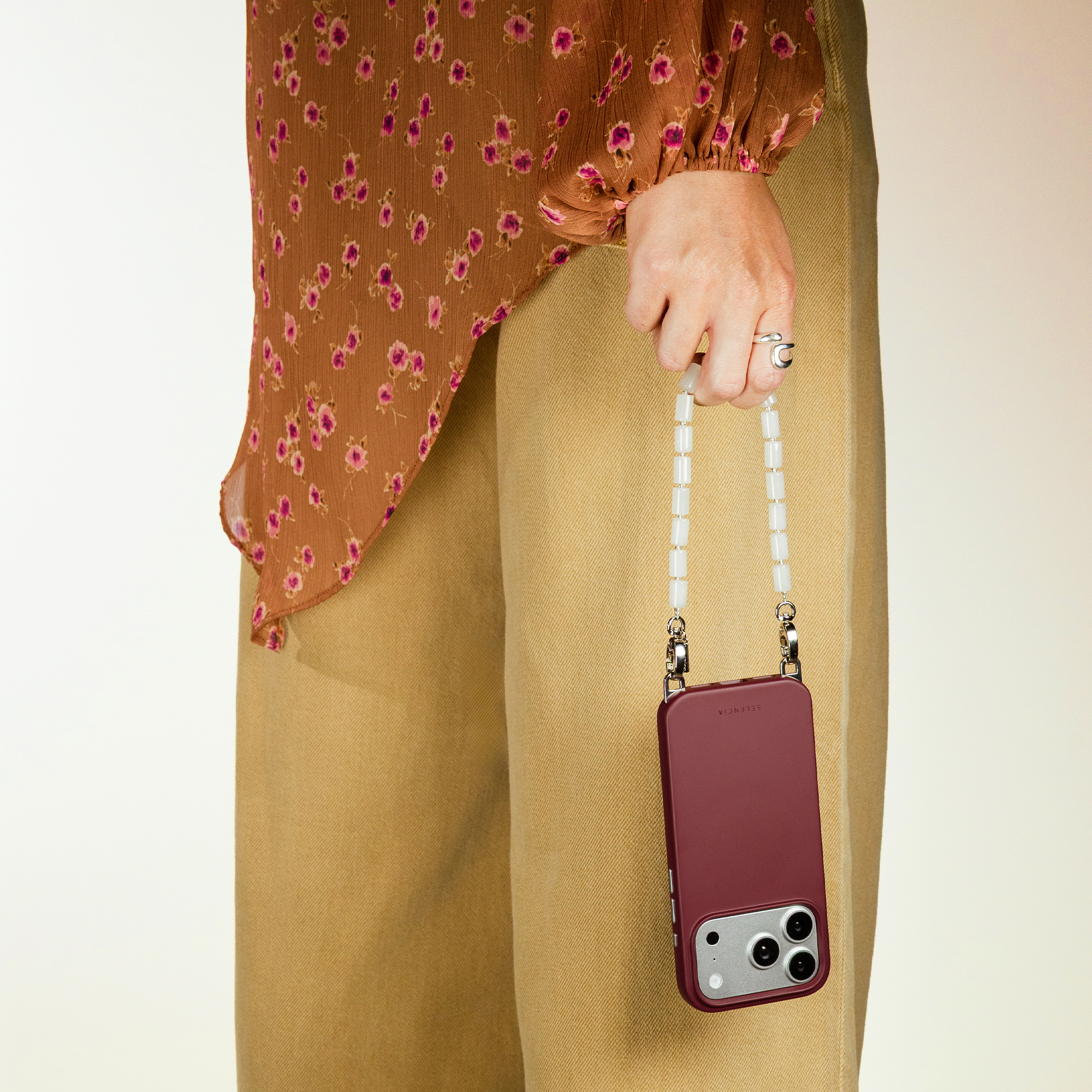 A person holds a burgundy phone case with a beaded strap. They wear a brown floral blouse.