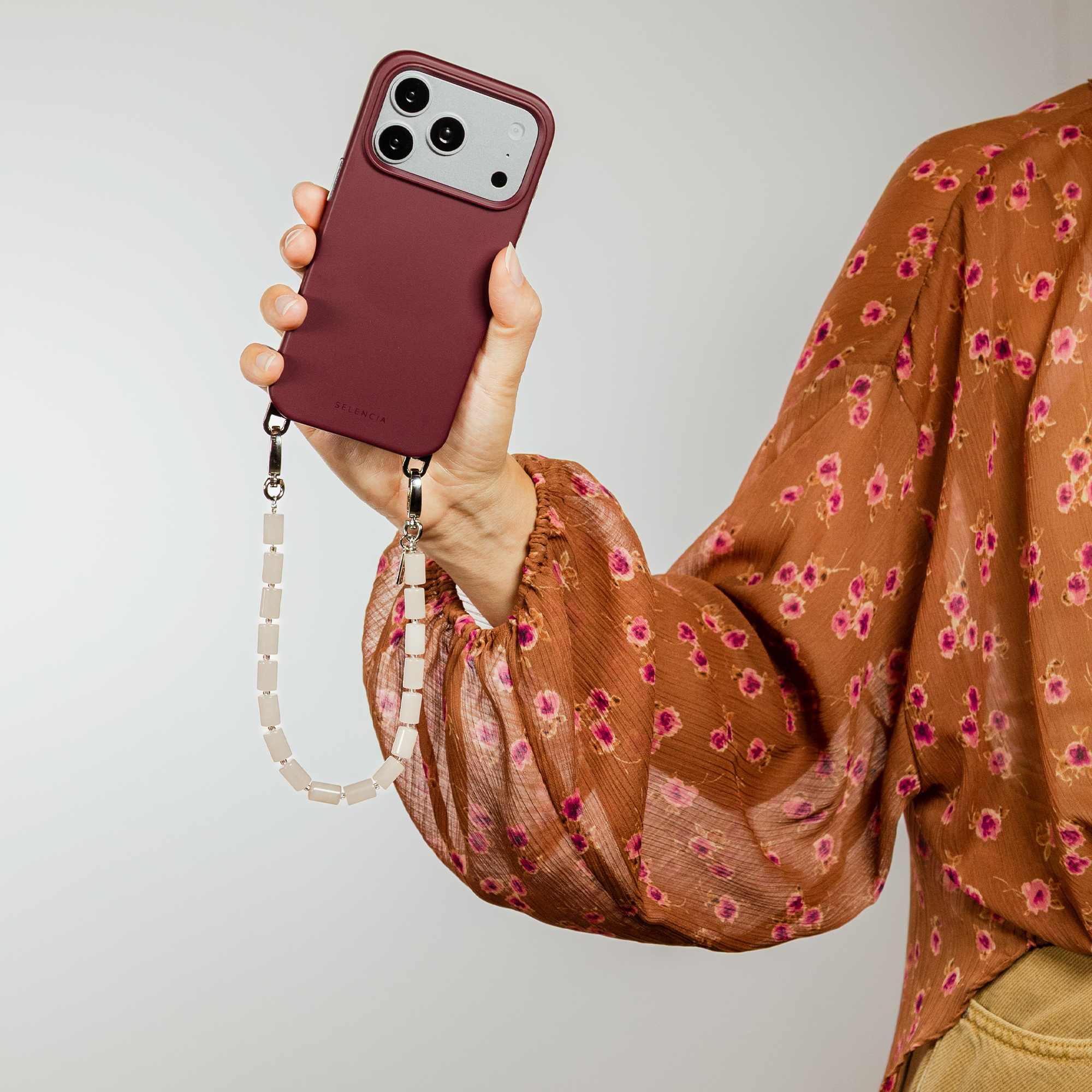 A hand holds a maroon phone with three cameras and a beaded strap.