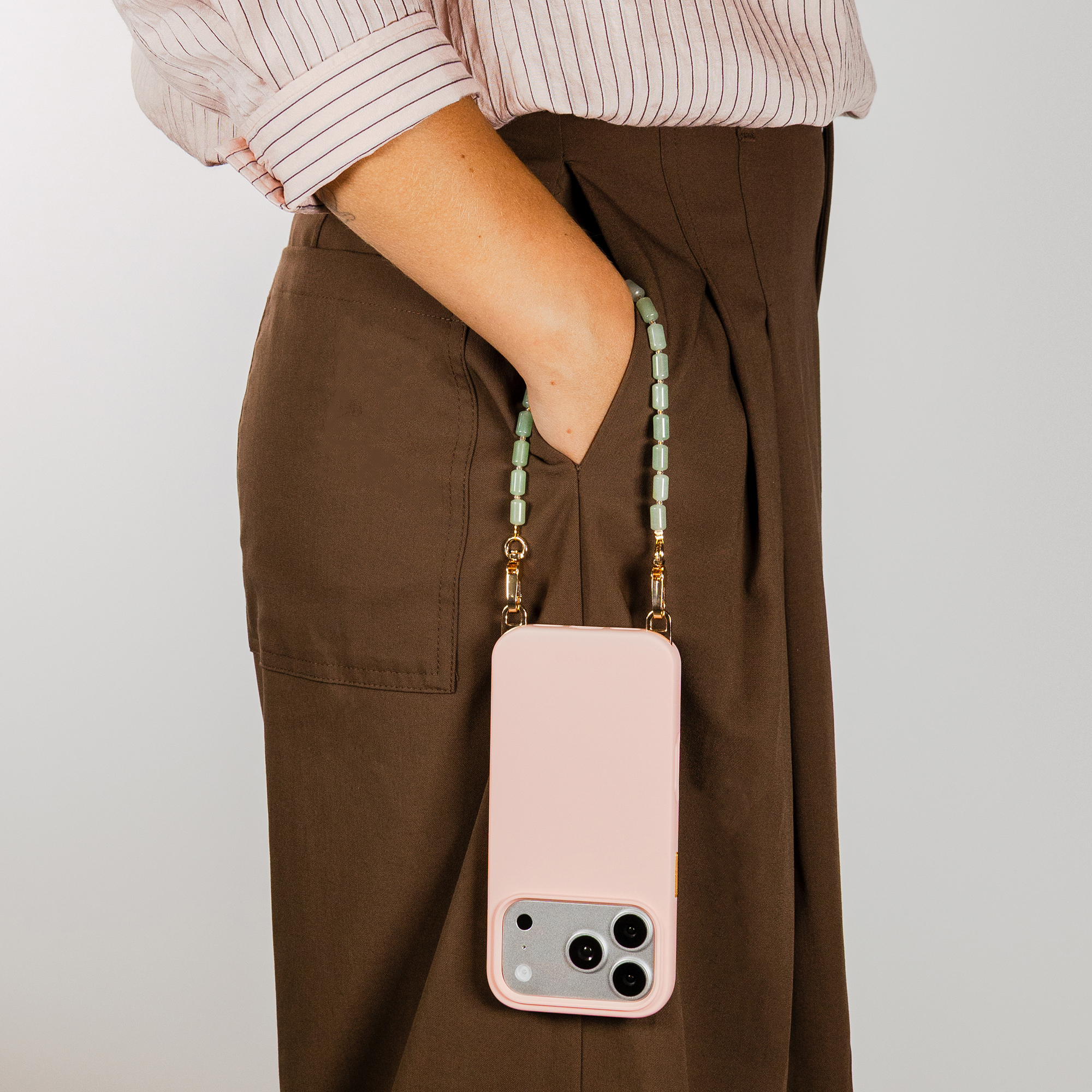 A person wearing a light pink striped blouse and brown pants holds a pink phone case on a beaded lanyard.