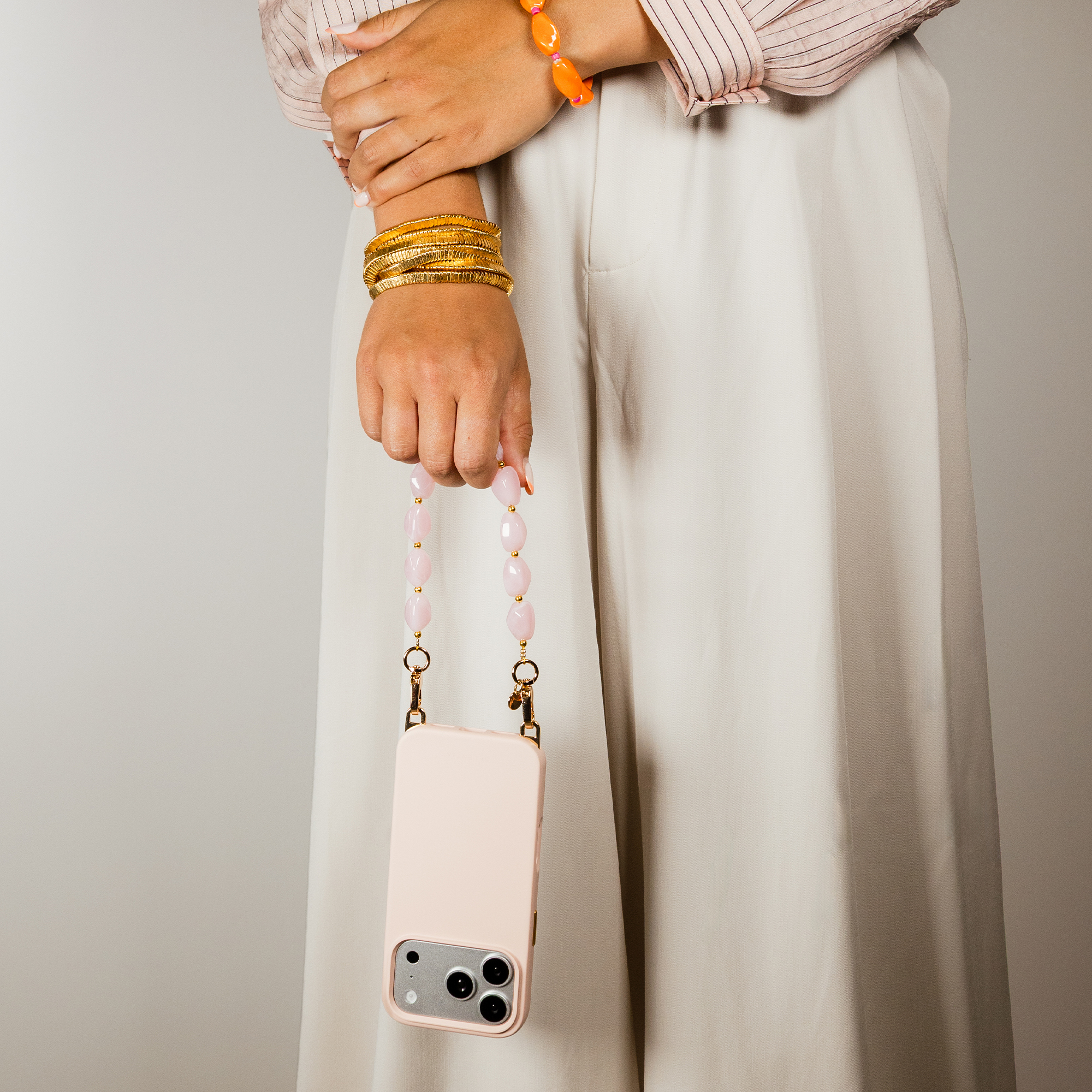 A person holds a light pink phone case with a beaded chain. Gold bangles.