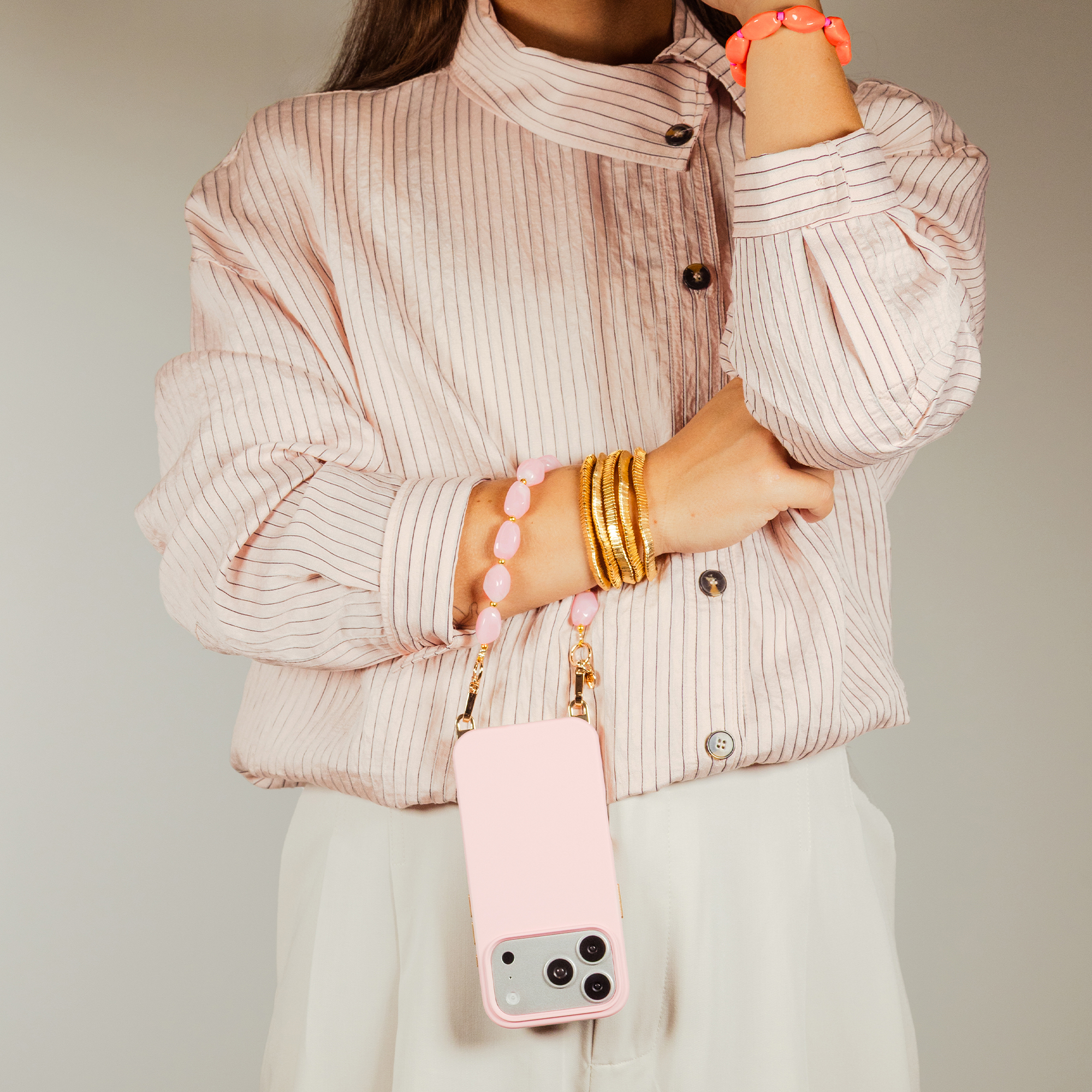 A person wears a striped pink shirt, white pants, gold bangles, and a pink phone case.