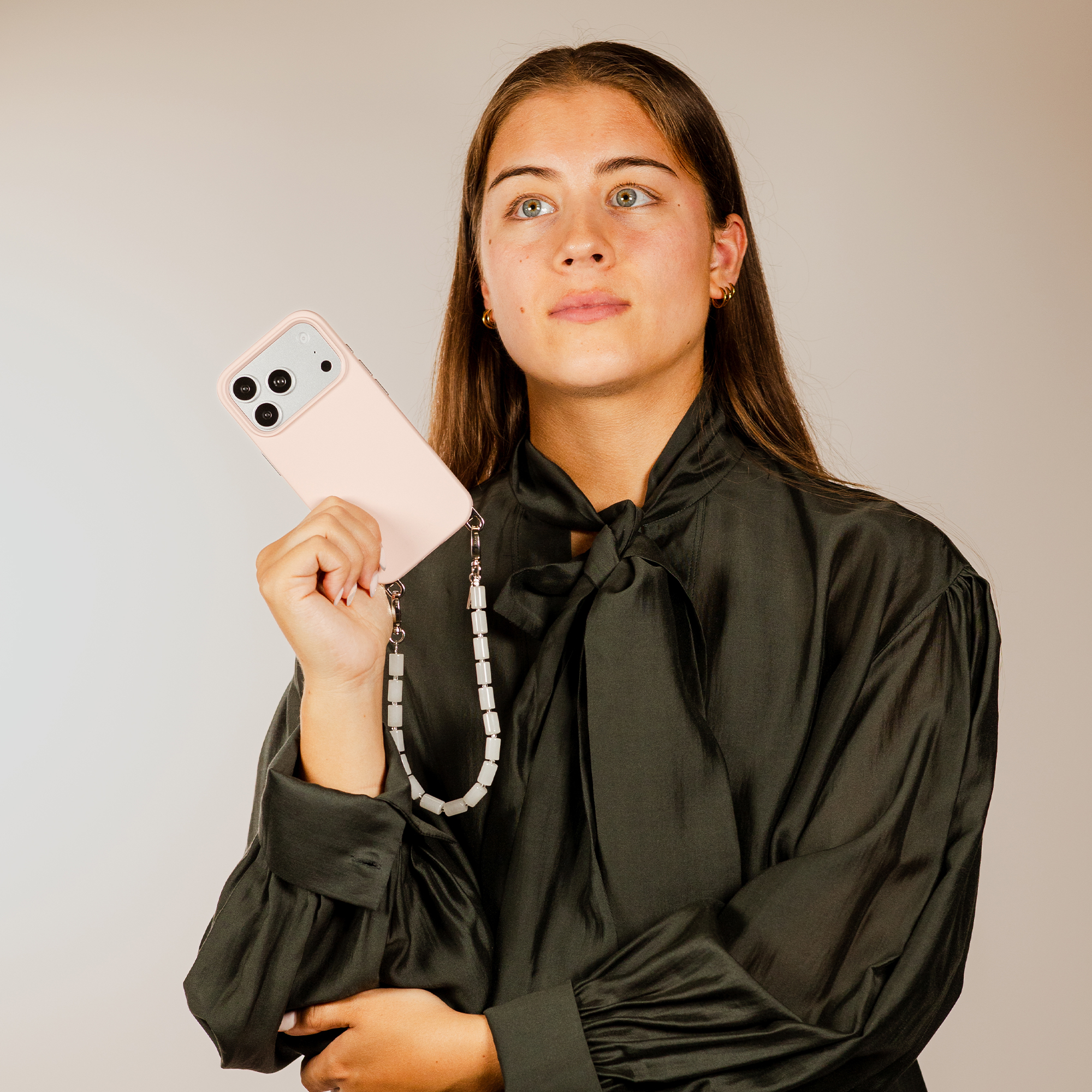 A young woman with brown hair holds a pink phone with a beaded strap.