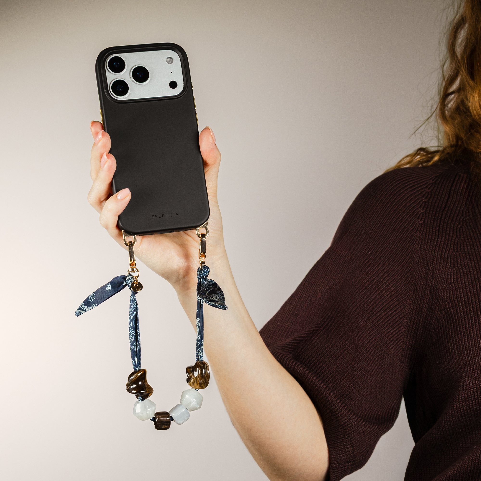 A person holds a black iPhone with a beaded lanyard and a paisley patterned ribbon.