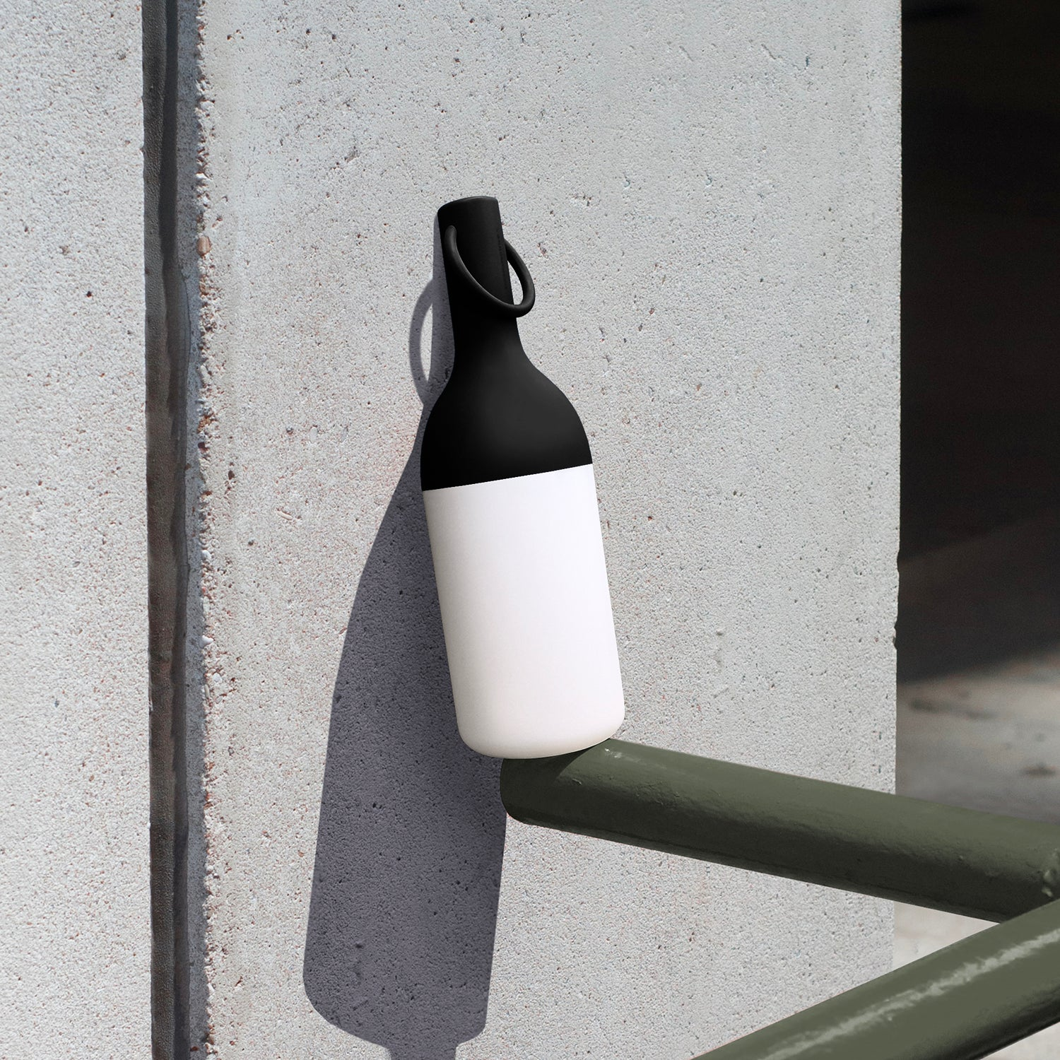 Schwarz-weiße Flasche mit Henkel ruht auf grünem Geländer vor Betonwand.
