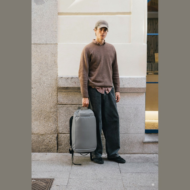 Uomo maglione marrone, camicia a quadri, jeans e cappellino tiene zaino grigio.