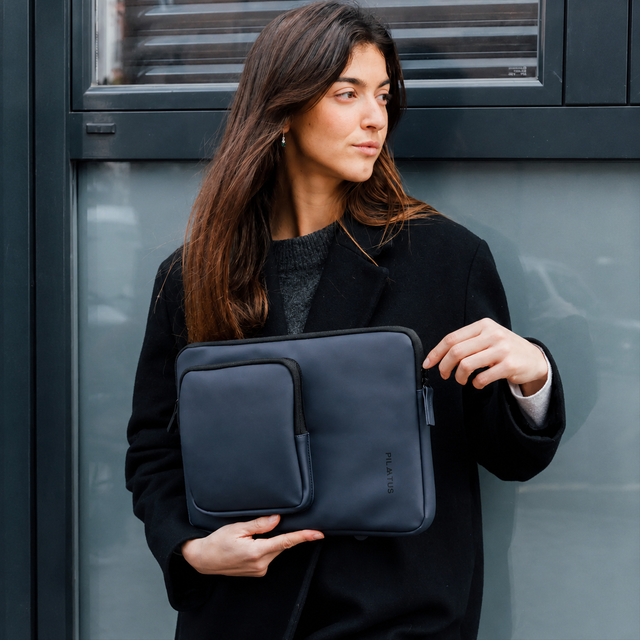 Una donna con i capelli scuri tiene una custodia per laptop blu scuro davanti a un muro grigio.