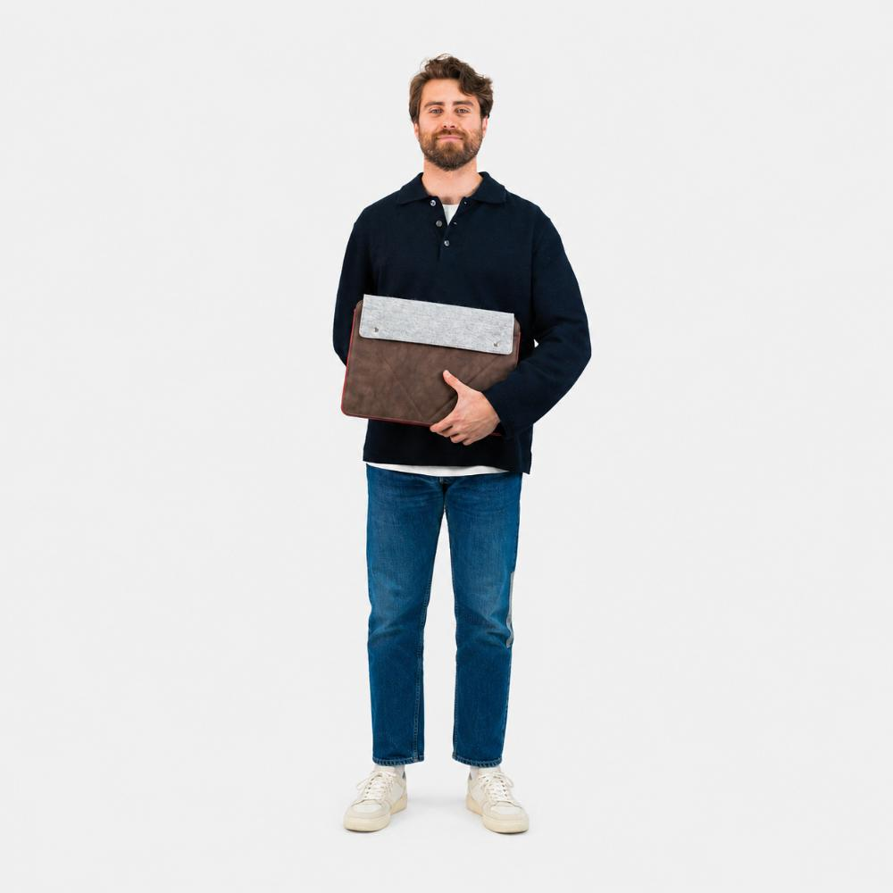 Uomo in maglione scuro e jeans tiene una custodia marrone per laptop con patta grigia.