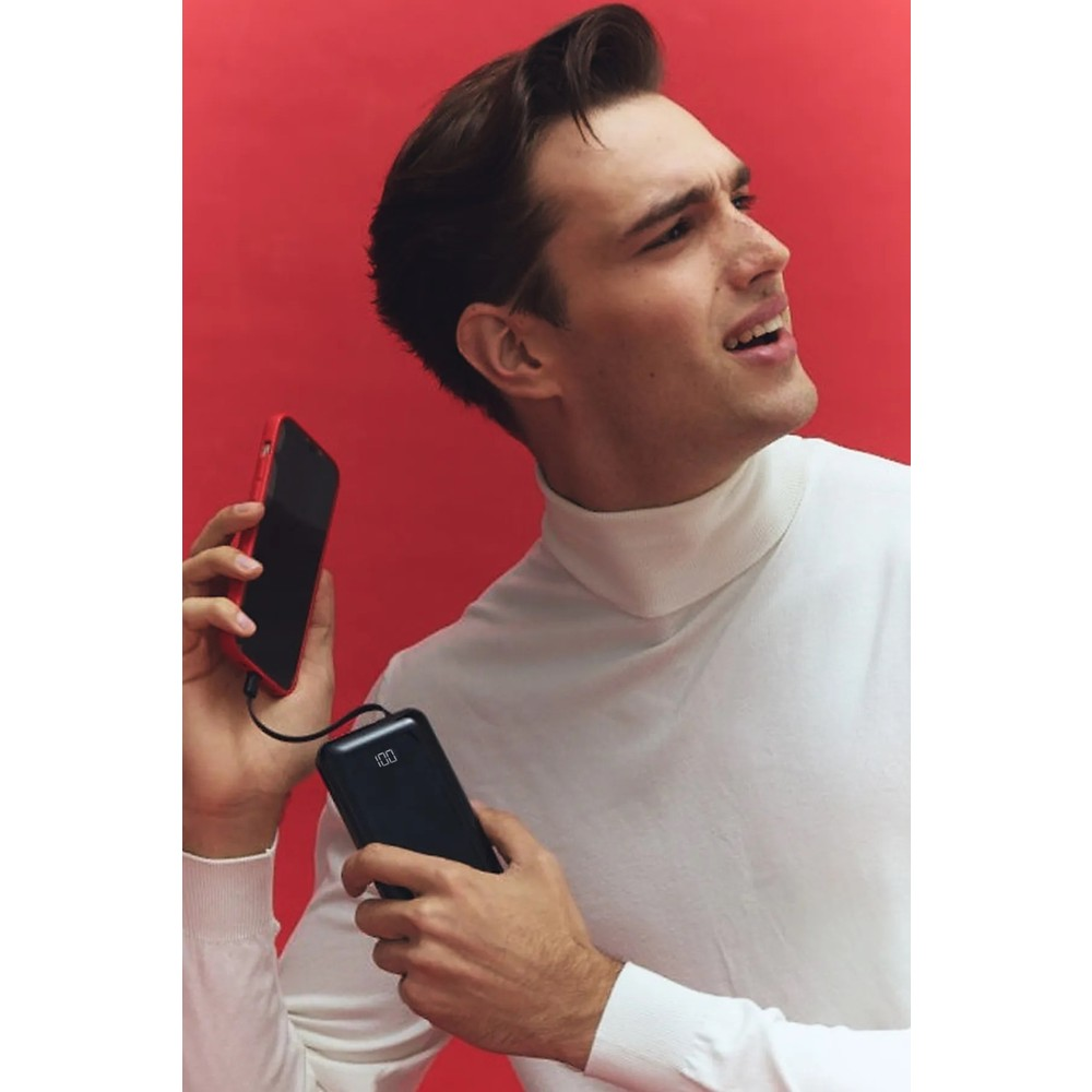 Beyaz balıkçı yaka kazaklı adam kırmızı telefon ve powerbank tutuyor.