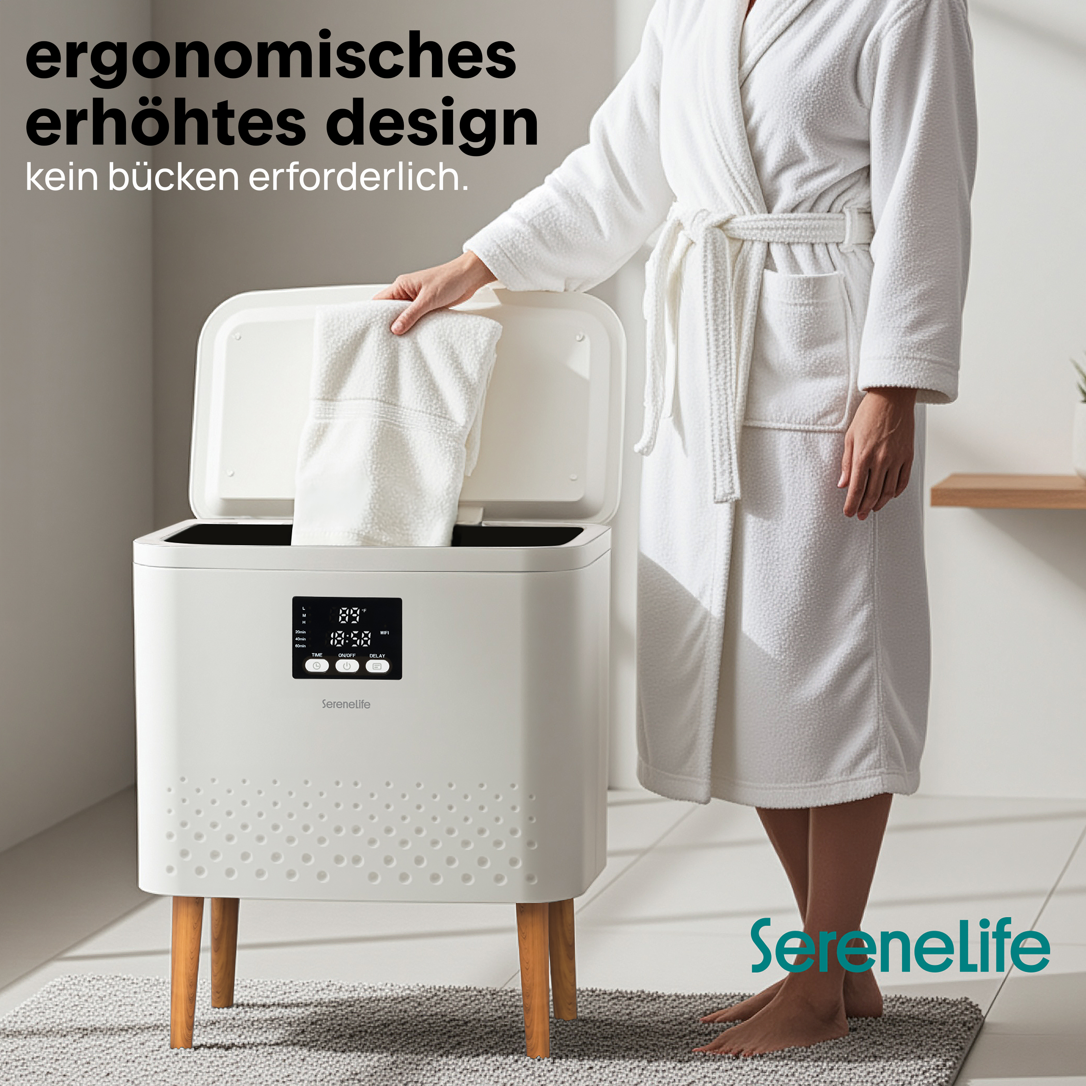 Eine Person im Bademantel legt ein Handtuch in ein weißes Gerät auf Holzbeinen.