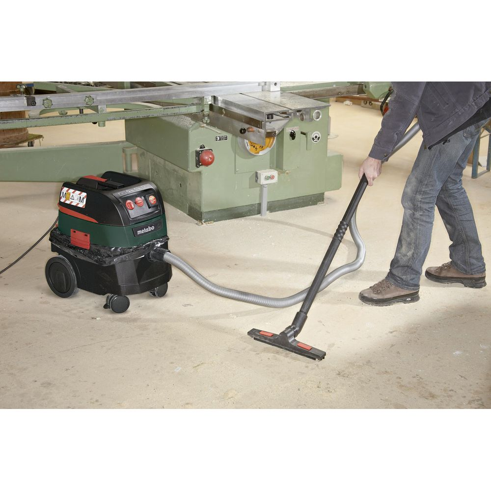 Un uomo aspira con un aspirapolvere Metabo, con una macchina da officina sullo sfondo.