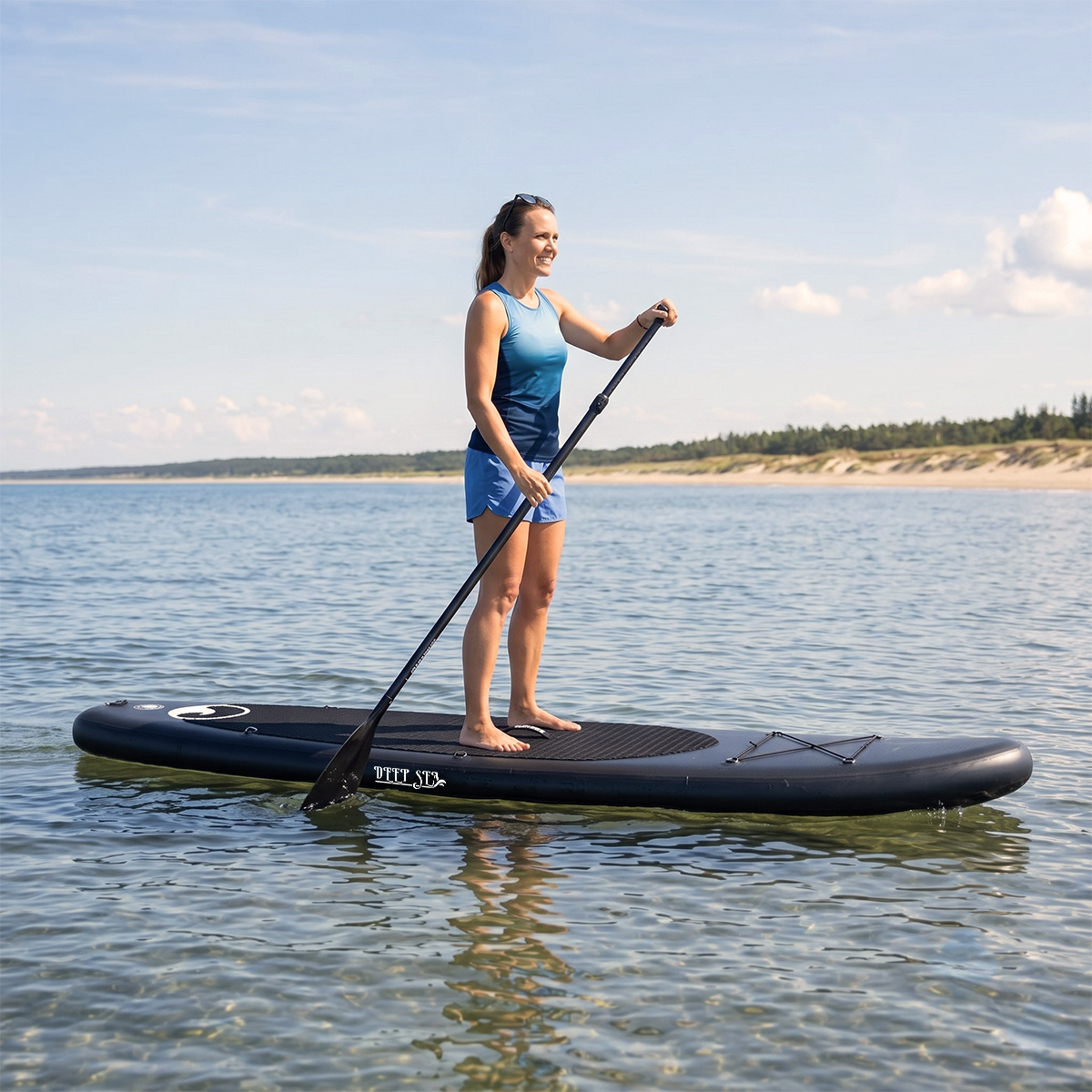 Eine Frau paddelt auf einem Board auf ruhigem Wasser vor einer sandigen Küste.