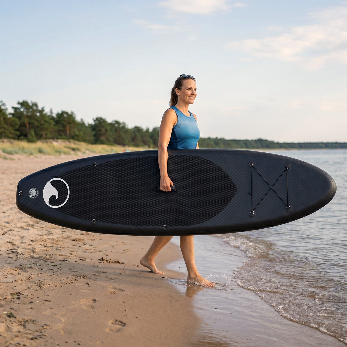Frau in blauem Oberteil trägt schwarzes Paddleboard am Strand. Wasser leckt Ufer.