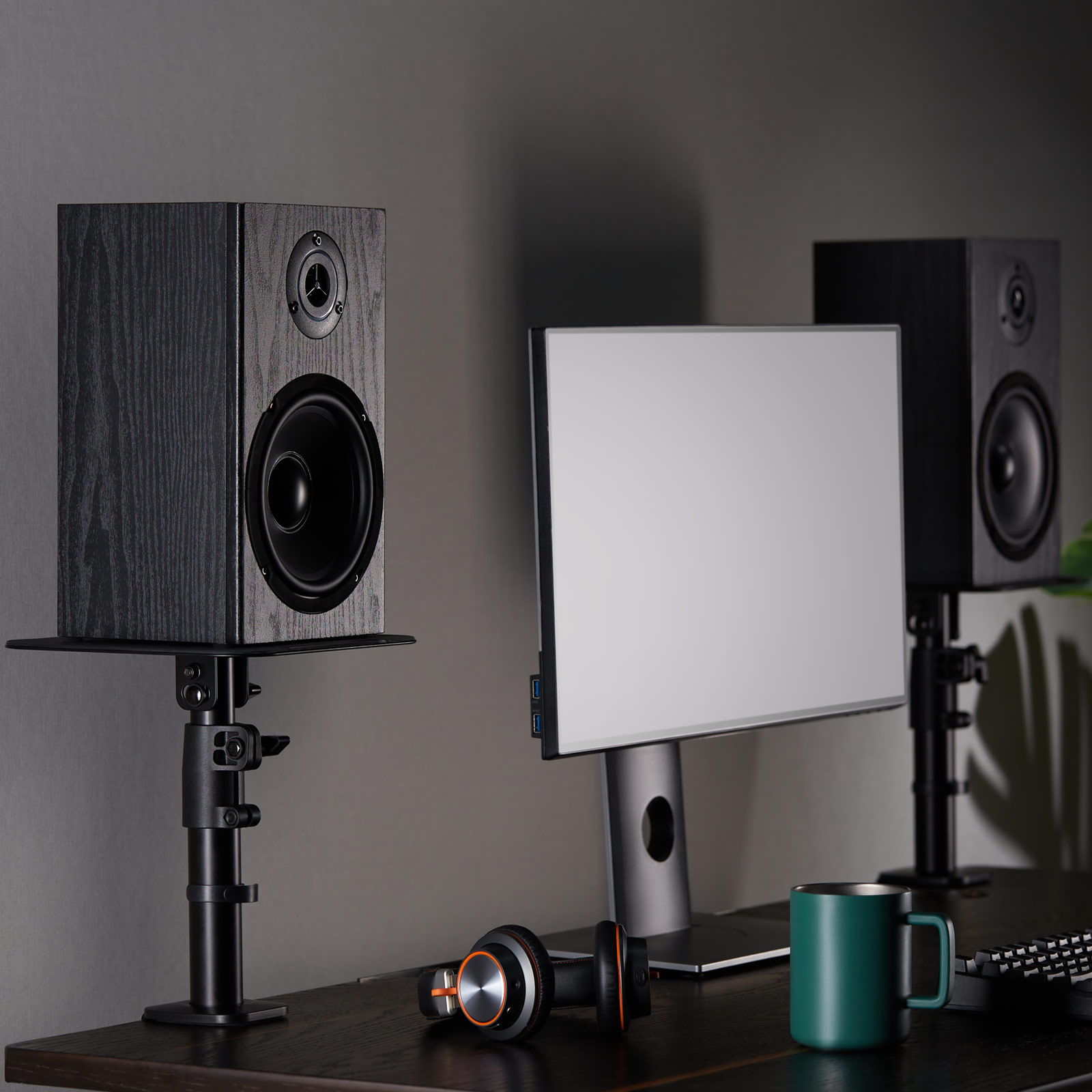Ein Schreibtisch-Setup mit zwei schwarzen Lautsprechern, einem Monitor, Kopfhörern und einer grünen Tasse.