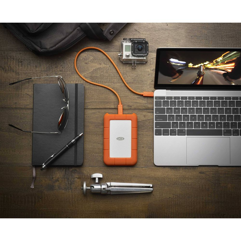 Laptop, disco esterno, GoPro, taccuino, occhiali da sole, penna e mini treppiede su legno.