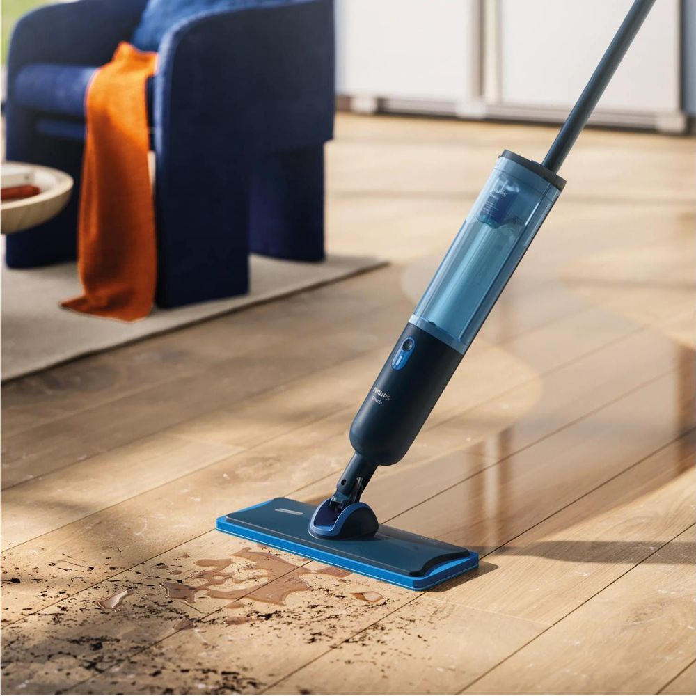 Un mocio spray blu pulisce un pavimento in legno sporco. Sullo sfondo una sedia blu con un panno arancione.