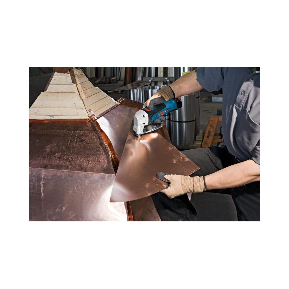 Handwerker schneidet Kupferblech mit Werkzeug. Innenraum mit Metallrollen im Hintergrund.