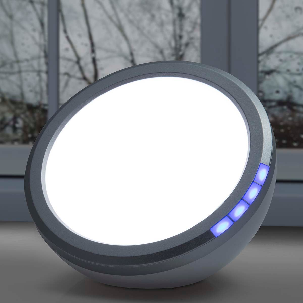 Ein ovaler, grauer Lichttherapie-Tisch mit blauen LEDs. Draußen ist es regnerisch.
