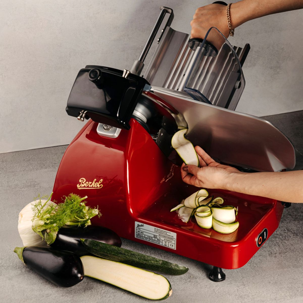 Una affettatrice Berkel rossa con zucchine, melanzane e finocchi. Una mano affetta zucchine.