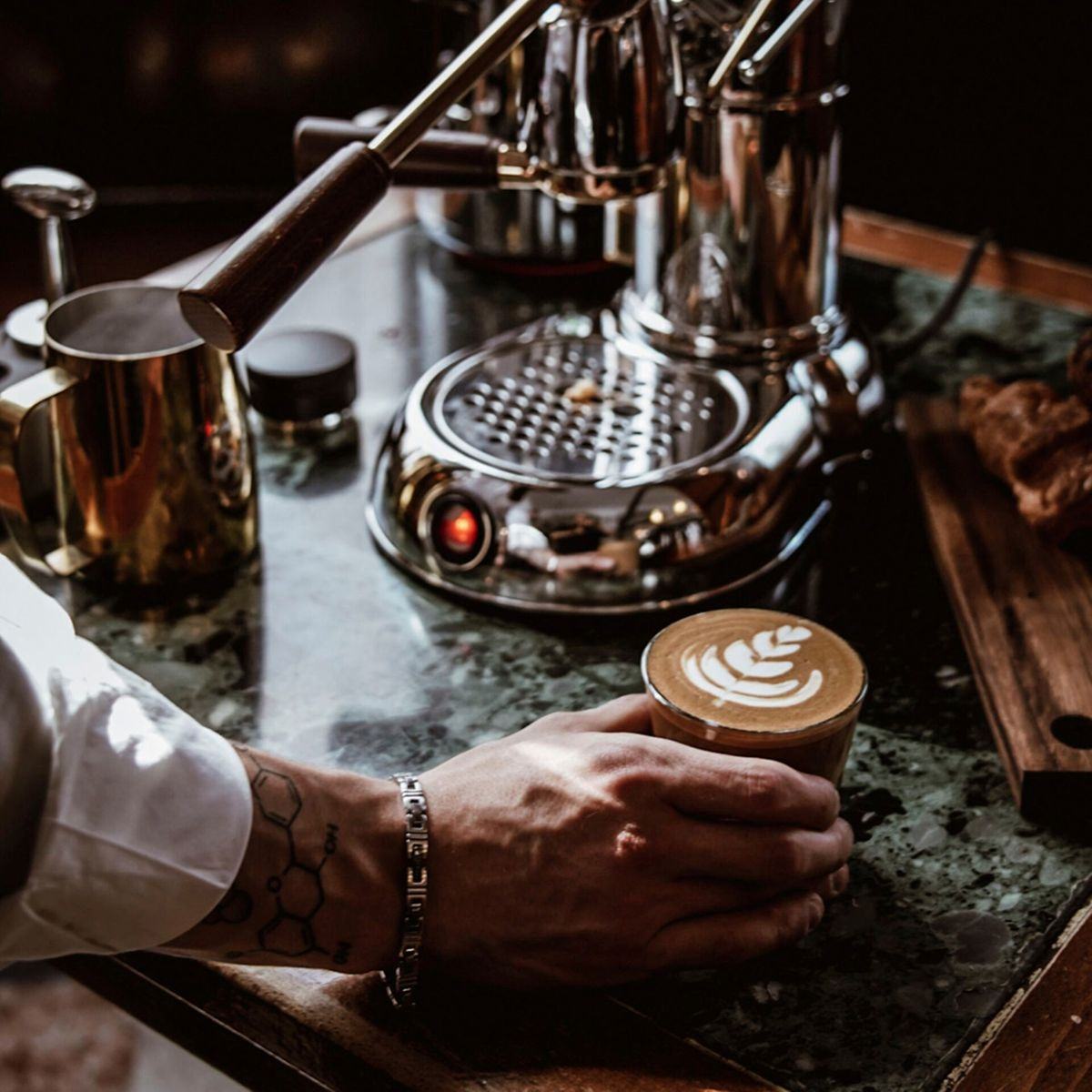 Osoba trzymająca filiżankę kawy latte art obok ekspresu do kawy.
