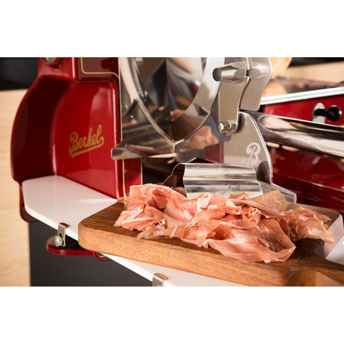 Prosciutto affettato sottile riposa su un tagliere di legno accanto a un'affettatrice Berkel rossa.