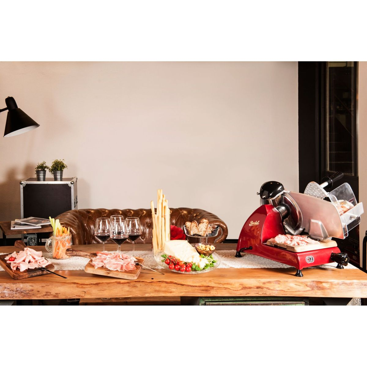 Affettatrice rossa su tavolo di legno con carne, pane e formaggio.