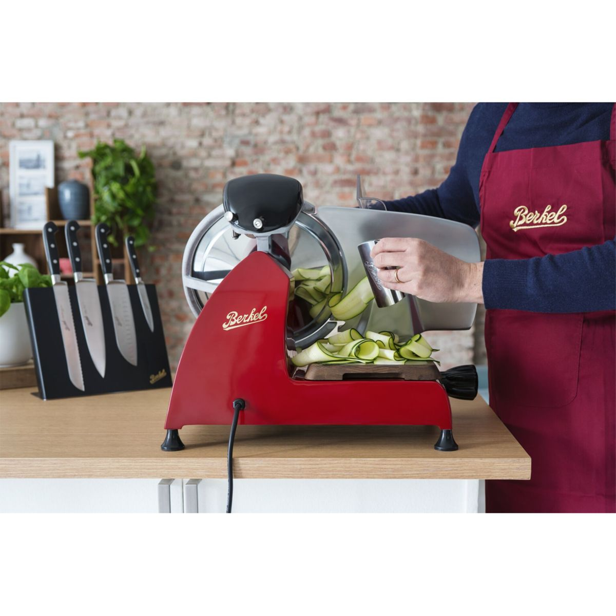 A person uses a red Berkel slicer to cut zucchini.