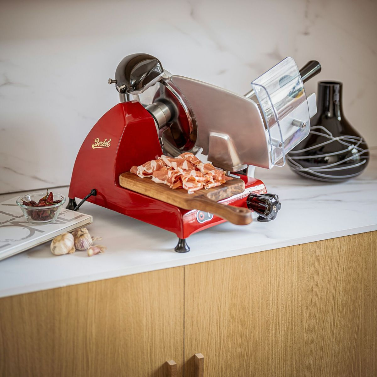 Red slicer with ham on a wooden board, beside garlic and chilies.