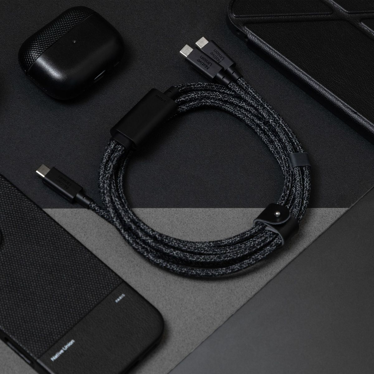 Schwarzes geflochtenes USB-C-Kabel mit Lederriemen, neben AirPods-Hülle und Handyhüllen.