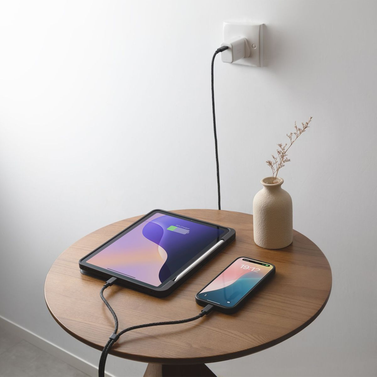 Tablet und Handy laden auf Holztisch neben Vase.