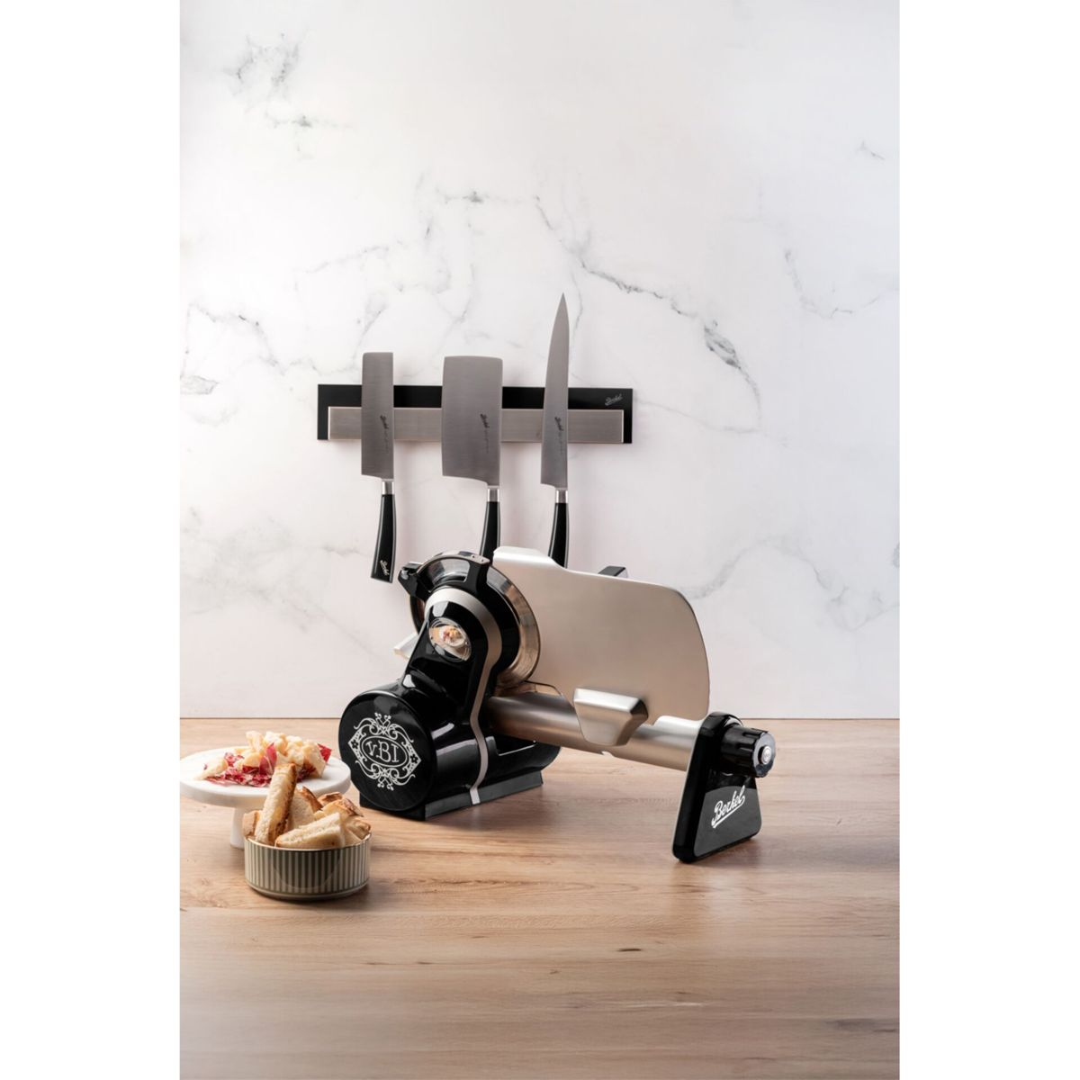 Affettatrice nera su tavolo in legno con pane e prosciutto. Coltelli appesi al muro.