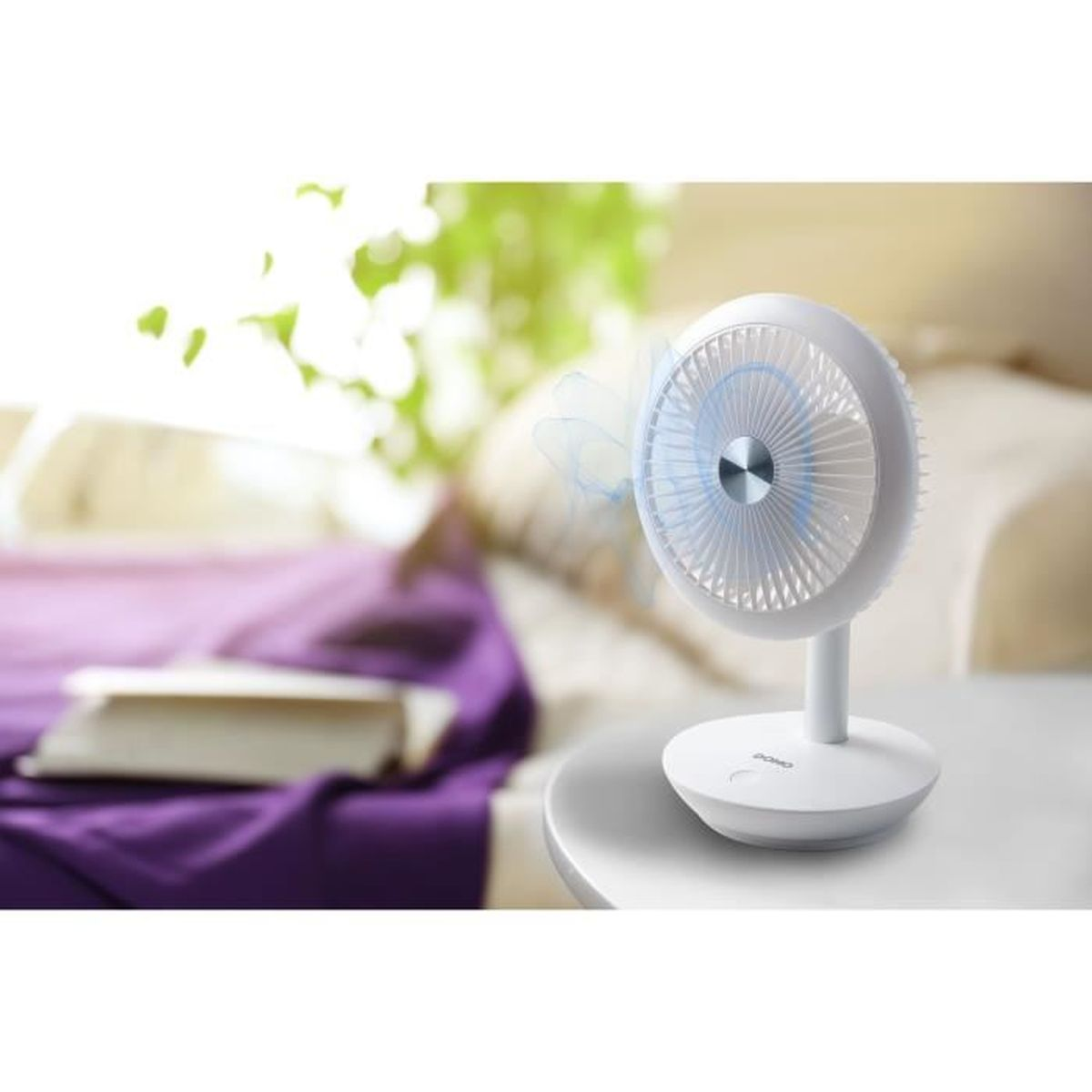 Ein weißer kleiner Ventilator steht auf einem Tisch. Im Hintergrund ein Bett mit lila Decke.