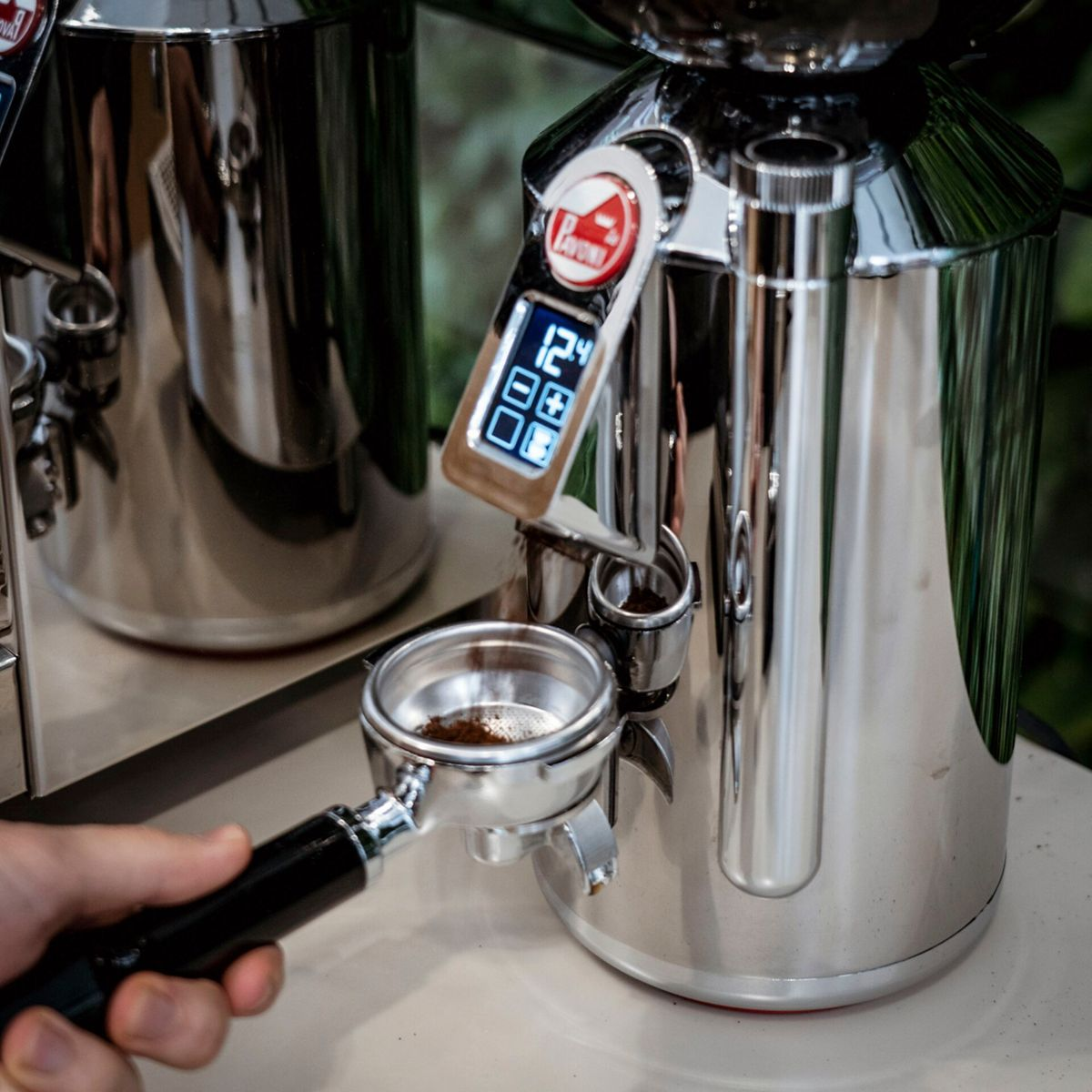 Eine Hand hält einen Siebträger unter eine Kaffeemühle mit digitaler Anzeige.